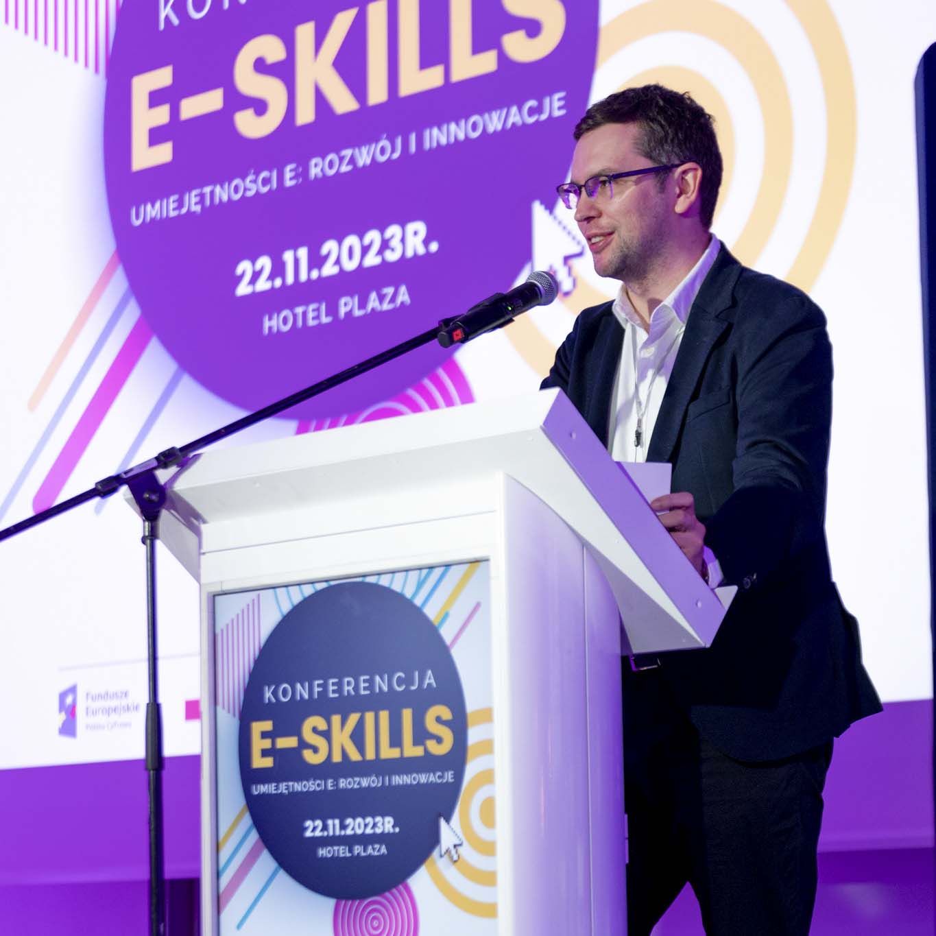 Przy mównicy stoi Pan Daniel Duda. Wzrok kieruje w stronę uczestników konferencji. Mówi do mikrofonu.