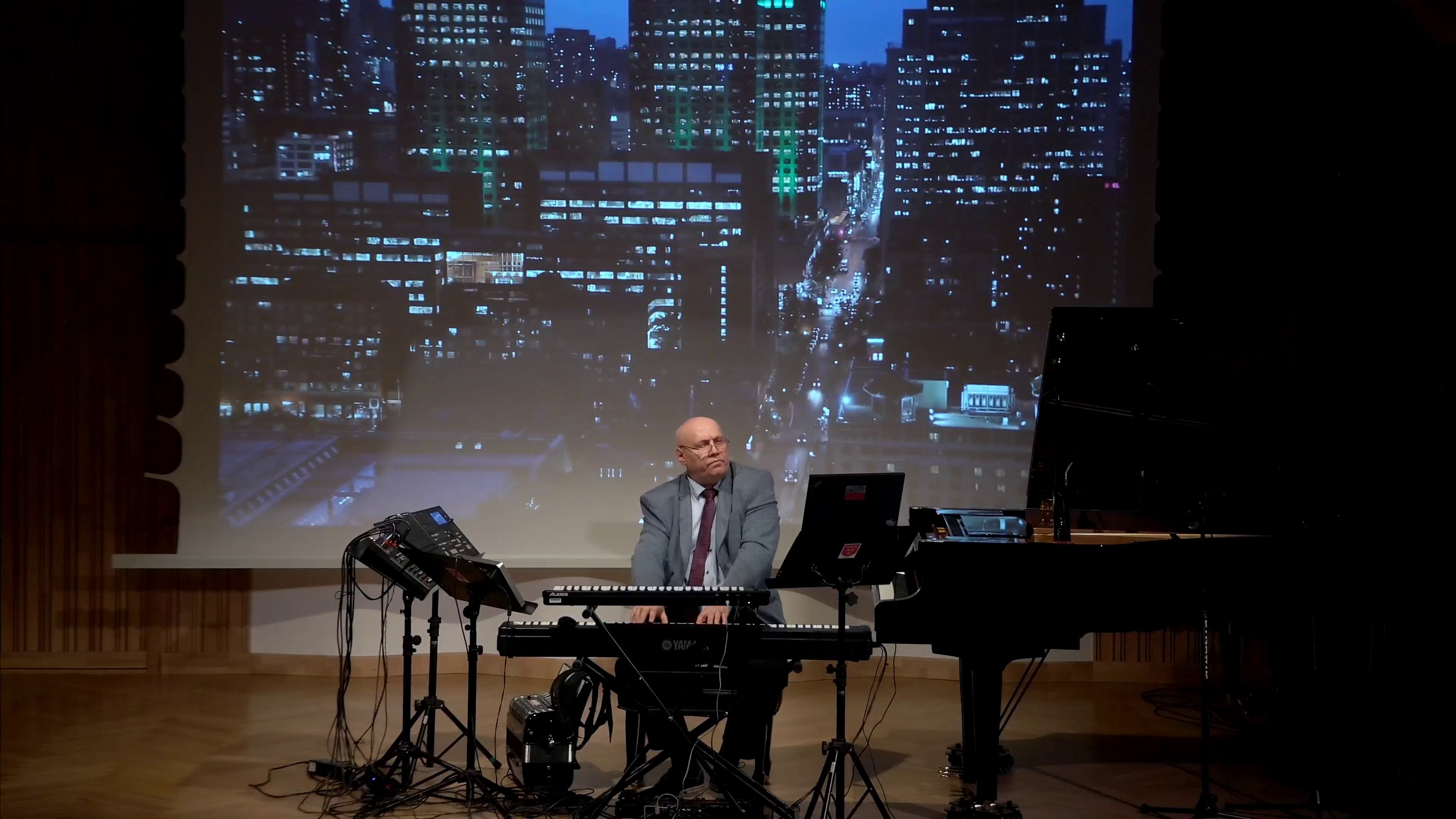 Zdjęcie. Estrada sali koncertowej. Na estradzie otwarty duży czarny fortepian oraz zestaw oplecionych gęstwiną kabli instrumentów elektronicznych, na których gra pan Dariusz Kownacki. W tel na ekranie wyświetlone jest zdjęcie miasta nocą.