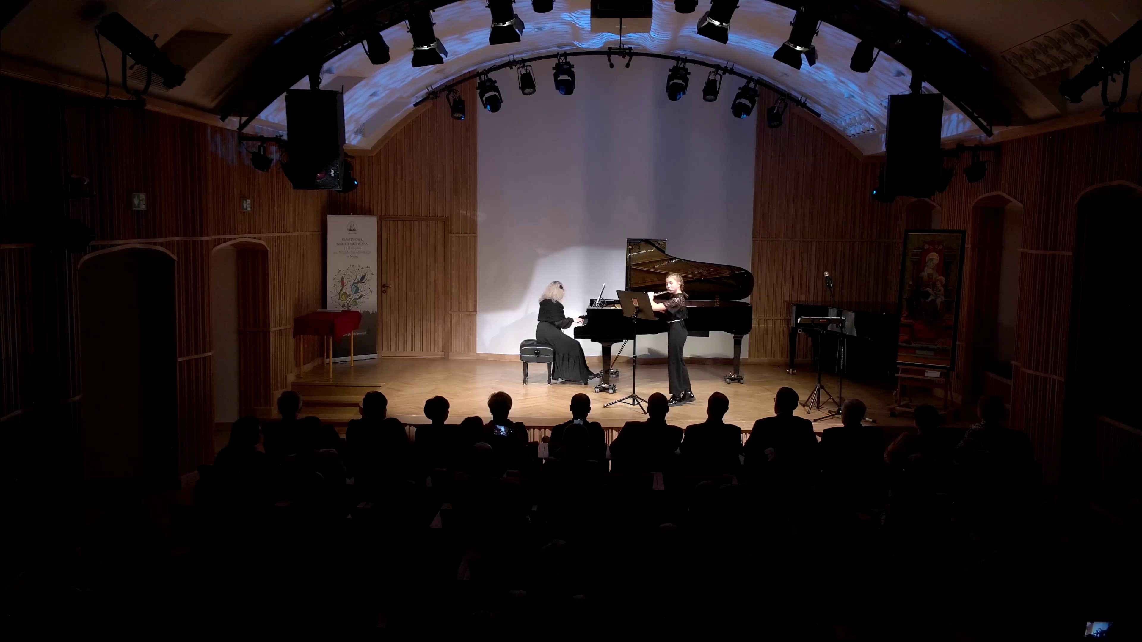 Zdjęcie. Na oświetlonej kameralnie estradzie gra młoda flecistka z towarzyszeniem pianistki, grającej na dużym czarnym fortepianie. Na pierwszym planie widać sylwetki wielu słuchaczy.