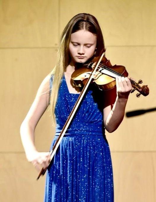 Zdjęcie przedstawia dziewczynkę w długich rozpuszczonych włosach, w niebieskiej sukience, która gra na skrzypcach. 