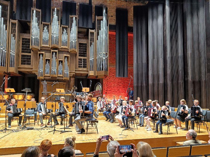 Uczniowie wraz z nauczycielami grający na akordeonach na scenie Filharmonii. W tle widoczne organy. Na dole zdjęcia widoczna widownia.