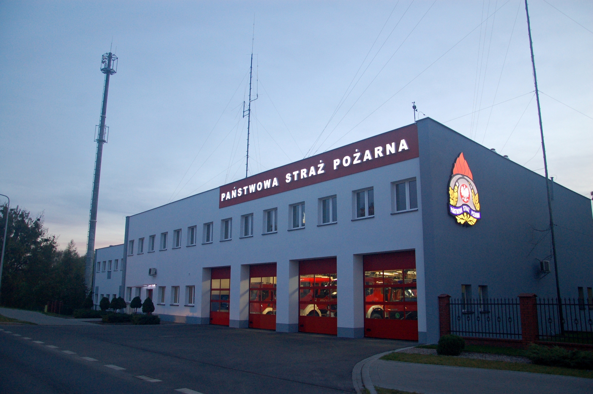 Zdjęcie przedstawia budynek Komendy Powiatowej PSP w Brodnicy od strony ulicy Sienkiewicza o zmierzchu. Na budynku podświetlony napis Państwowa Straż Pożarna oraz logo PSP. W przeszklonych wrotach garażowych widoczny oświetlone pojazdy Jednostki Ratowniczo-Gaśniczej.
