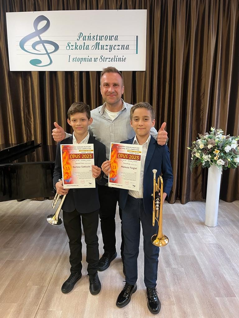 Na zdjęciu widoczny jest nauczyciel Paweł Ficoń oraz dwoje uczniów trzymających dyplomy konkursowe oraz trąbki. Wszyscy mają na sobie eleganckie stroje. 