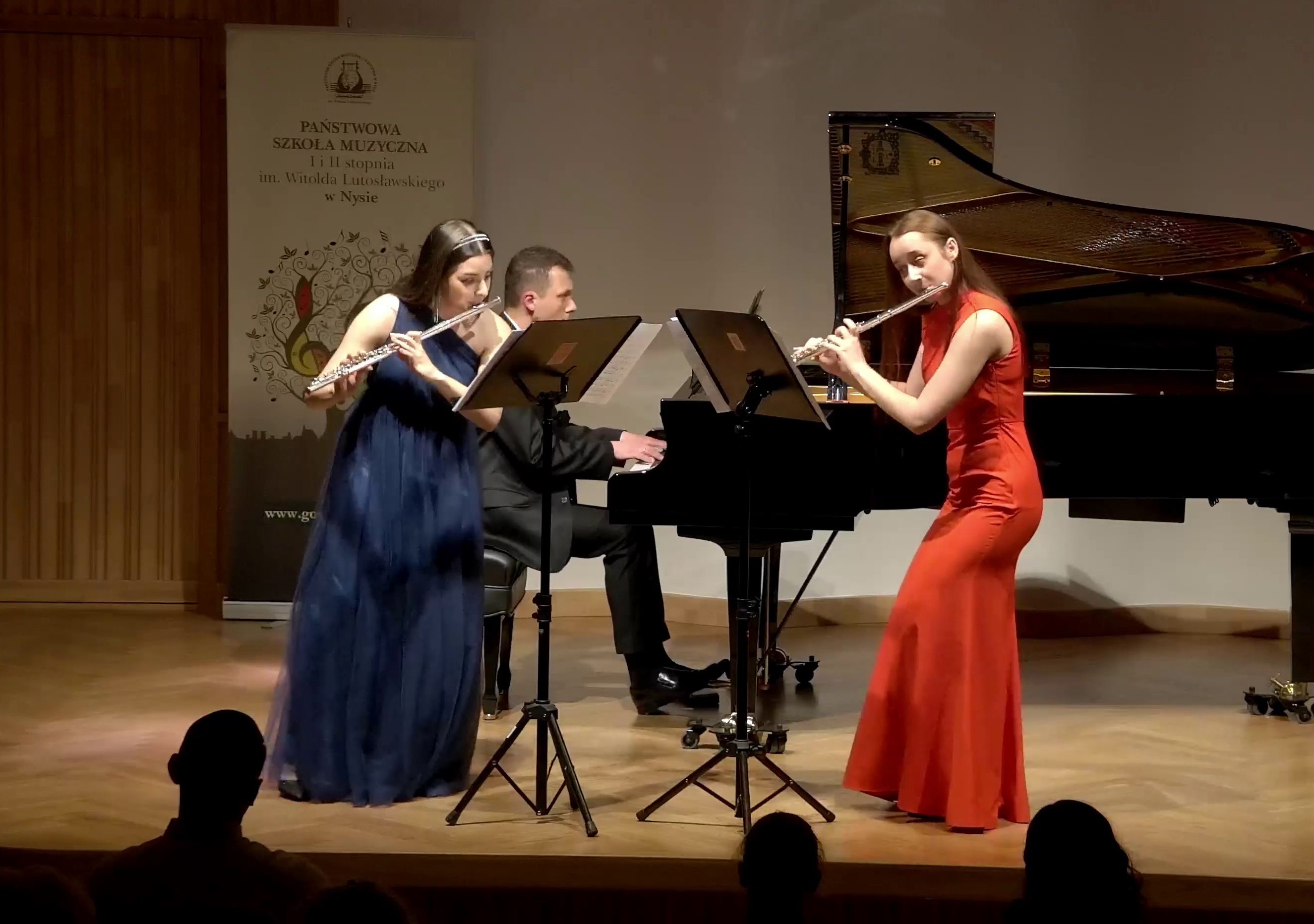 Na estradzie występują dwie flecistki z towarzyszeniem pianisty. Prze flecistkami stoją pulpity z nutami. W tle czarny fortepian oraz baner PSM Nysa
