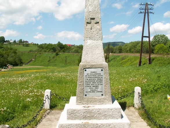 Pomnik upamiętniający miejsce egzekucji na nim tablica i krzyż. W tle zieleń trawy oraz linia kolejowa.