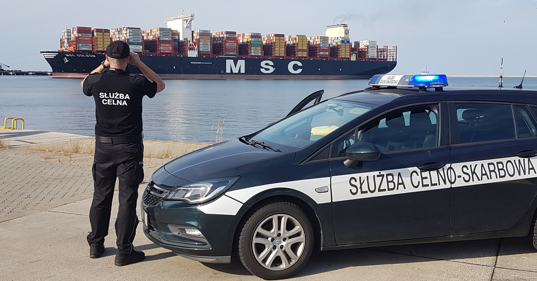 Funkcjonariusz Służby Celno-Skarbowej stojąc przy samochodzie obserwuje przez lornetkę kontenerowiec
