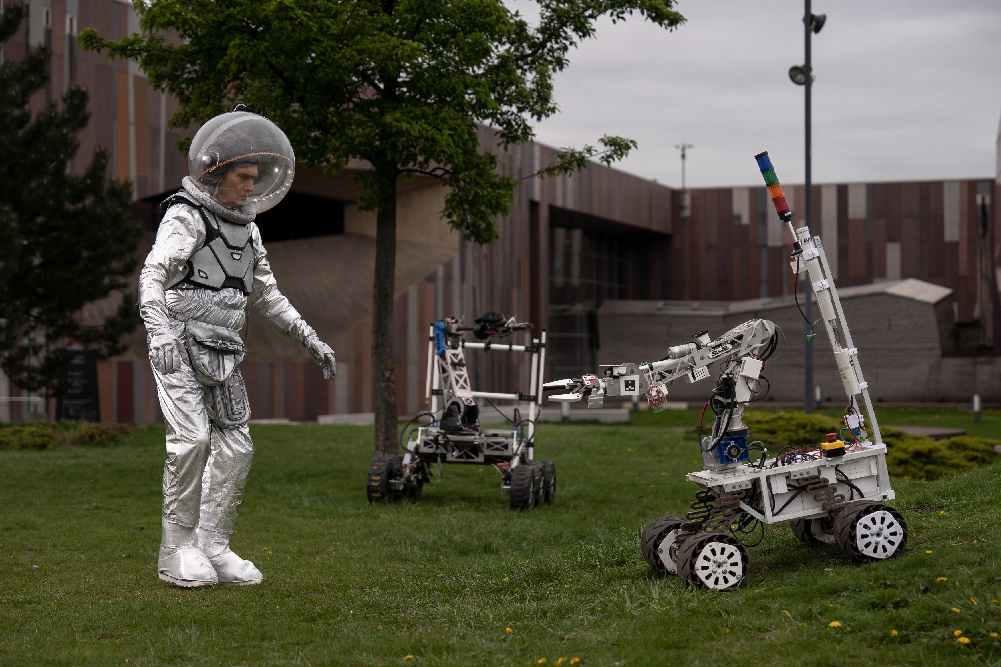 Mężczyzna w stroju astronauty stoi na trawniku przed budynkiem, przed nim dwa łaziki