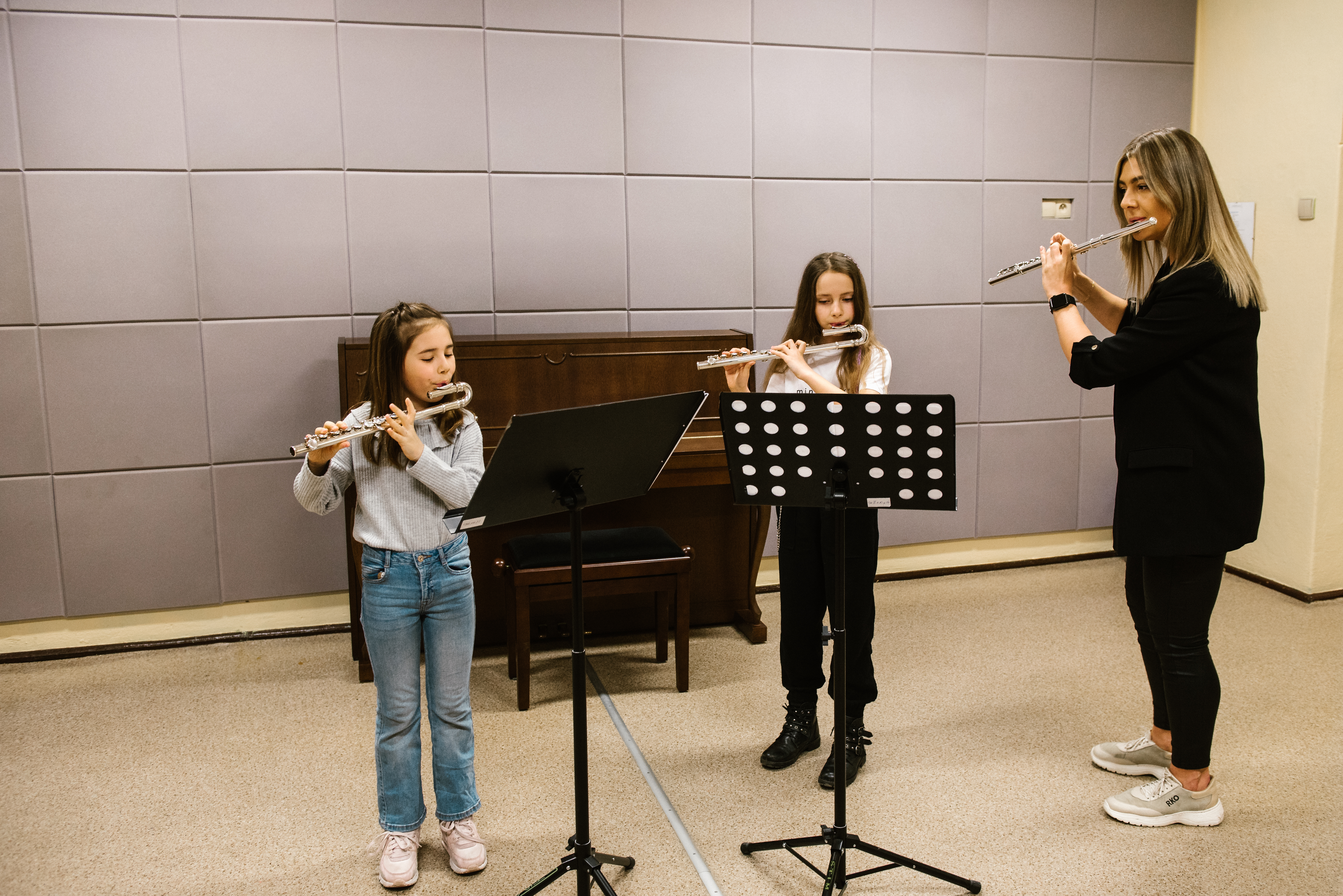 Na zdjęciu znajduje się nauczycielka Agata Podlejska oraz uczennice Alicja Sobczak i Liliana Bisikiewicz grające na fletach z nut z czarnych pulpitów. W tle stoi brązowe pianino. 