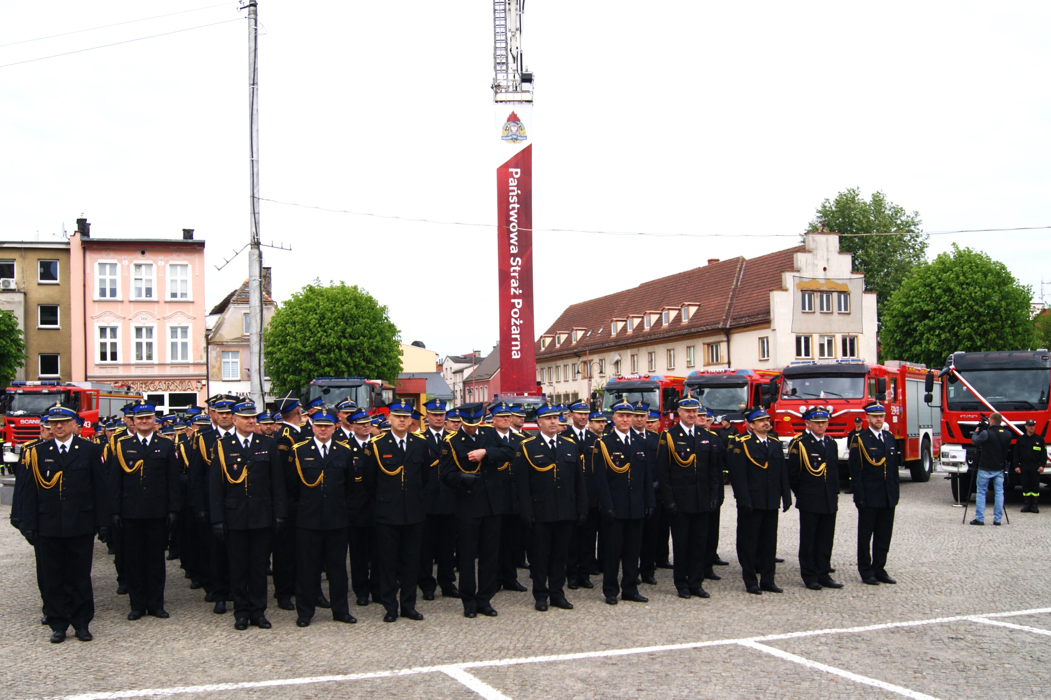 Na placu miejskim stoi kompania strażaków w mundurach galowych