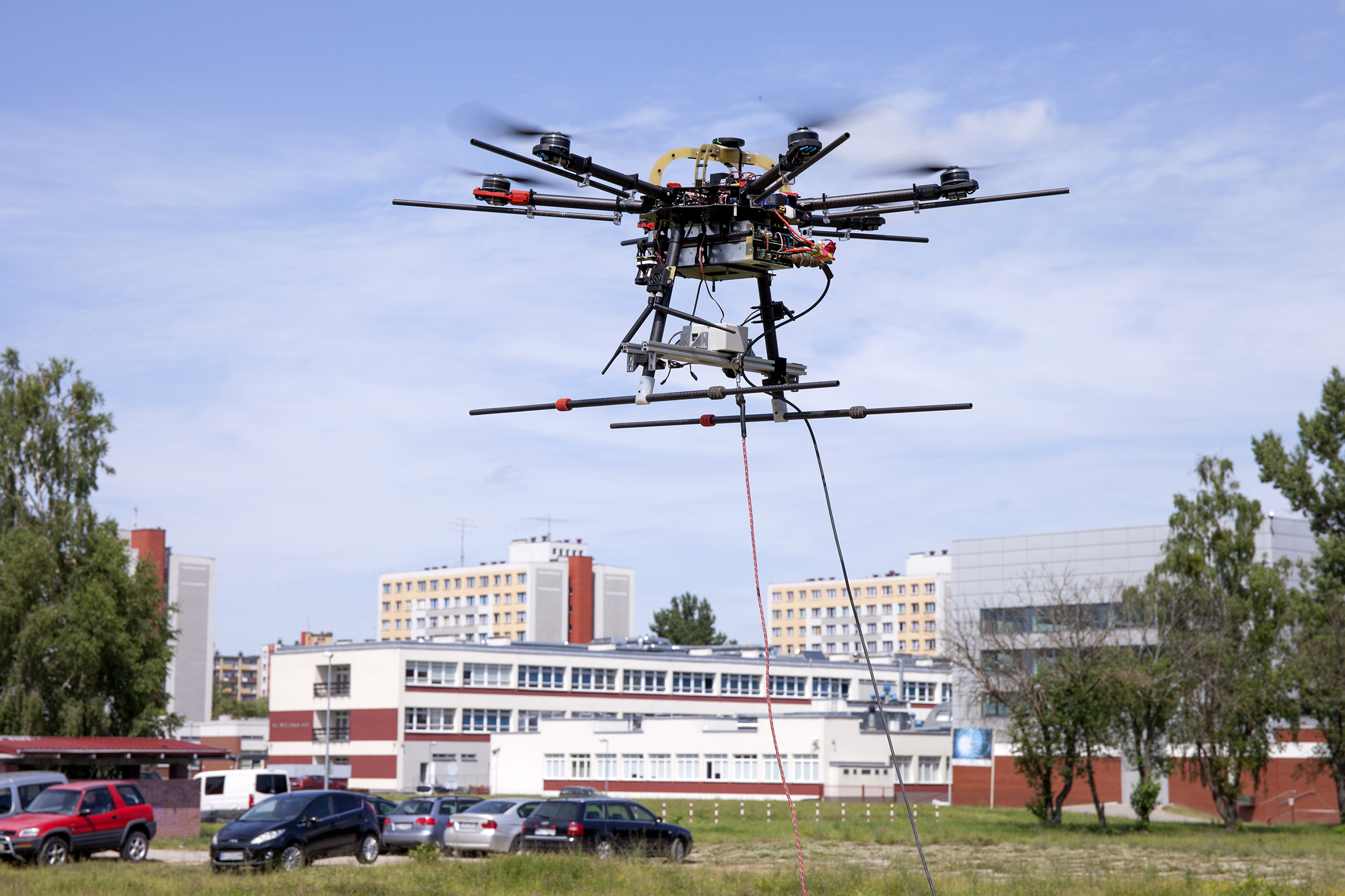 Testy elementów systemu AVAL przeprowadzone na Politechnice Białostockiej. W centrum zdjęcia widoczny dron wznoszący się nad łąką i parkingiem w pobliżu kampusu uczelni