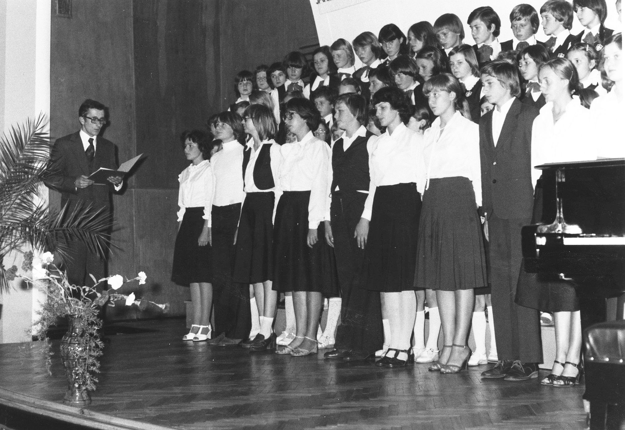 Zdjęcie. Inauguracja działalności Szkoły II stopnia – ślubowanie uczniów odbiera dyrektor Stanisław Cetnarowski. Wszyscy stoją na estradzie w sali koncertowej szkoły. Przed nim w lewym dolnym rogu wazon z kwiatami - goździkami i fragment palmy. Dyrektor Cetnarowski odczytuje tekst ślubowania. Uczniowie ustawieni sa na schodkowych podestach - jak chór - i wsłuchują się w słowa ślubowania.