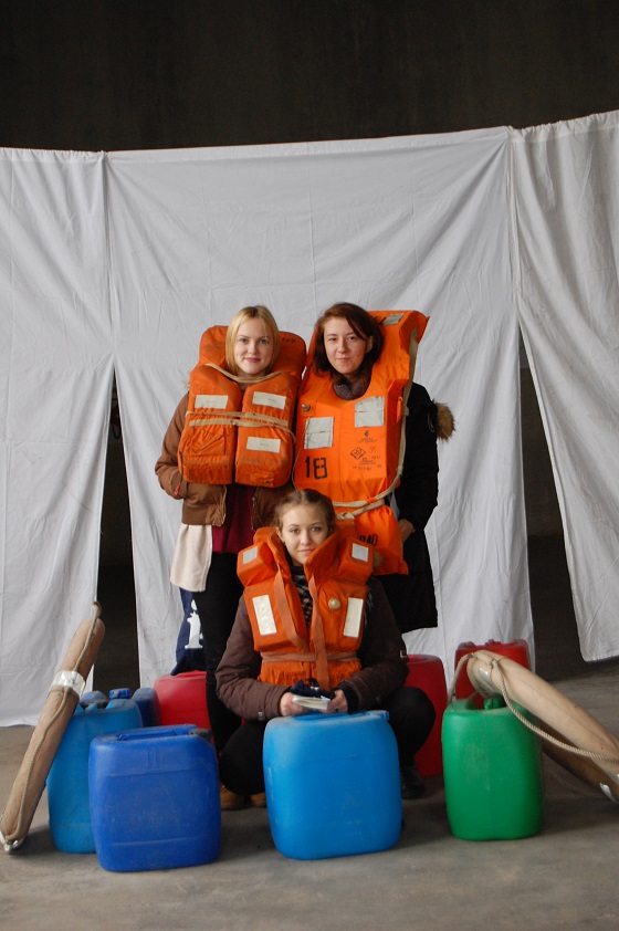 Three schoolgirls dressed in life jackets pose. Cans for a water and lifebuoys are around them. 