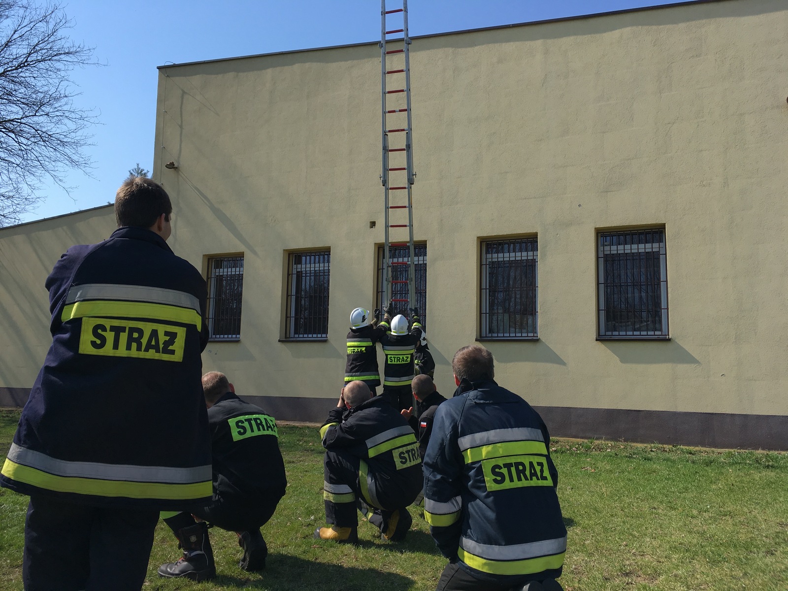 Zdjęcie przedstawia grupę strażaków OSP w trakcie przystawiania drabiny pożarniczej do budynku.