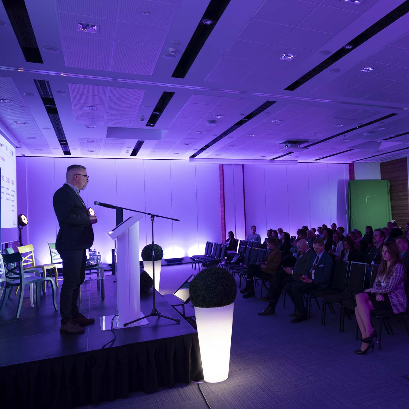 Ujęcie z boku sceny. Przy mównicy stoi Pan Norbert Ogłaza, naprzeciwko niego siedzą uczestnicy konferencji.