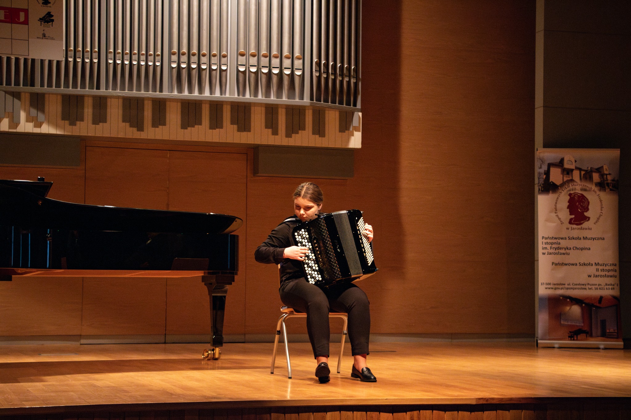 Na drewnianej scenie, przed ścianą z drewnianą boazerią i dużymi organami piszczałkowymi, siedzi młoda kobieta grająca na akordeonie. Kobieta ma ciemne włosy spięte w kok i ubrana jest w ciemną bluzkę z długimi rękawami oraz ciemne spodnie. Siedzi na metalowym krześle z okrągłym siedziskiem, a jej stopy spoczywają płasko na podłodze. Akordeon, na którym gra, jest czarny z białymi i czarnymi klawiszami. Jej dłonie są ułożone na klawiszach i guzikach instrumentu. Po lewej stronie sceny stoi czarny fortepian ze otwartą klapą. W tle, po prawej stronie, widoczny jest pionowy baner z tekstem w języku polskim i wizerunkiem Fryderyka Chopina w medalionie. Oświetlenie sceniczne jest skupione na grającej kobiecie, tworząc jaśniejszy obszar wokół niej na tle ciemniejszej reszty sceny i ściany.