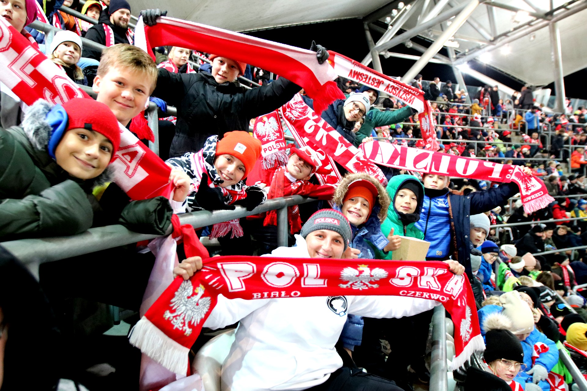Duża grupa roześmianych dzieci siedzi na trybunie stadionu, machają biało-czerwonymi szalikami.