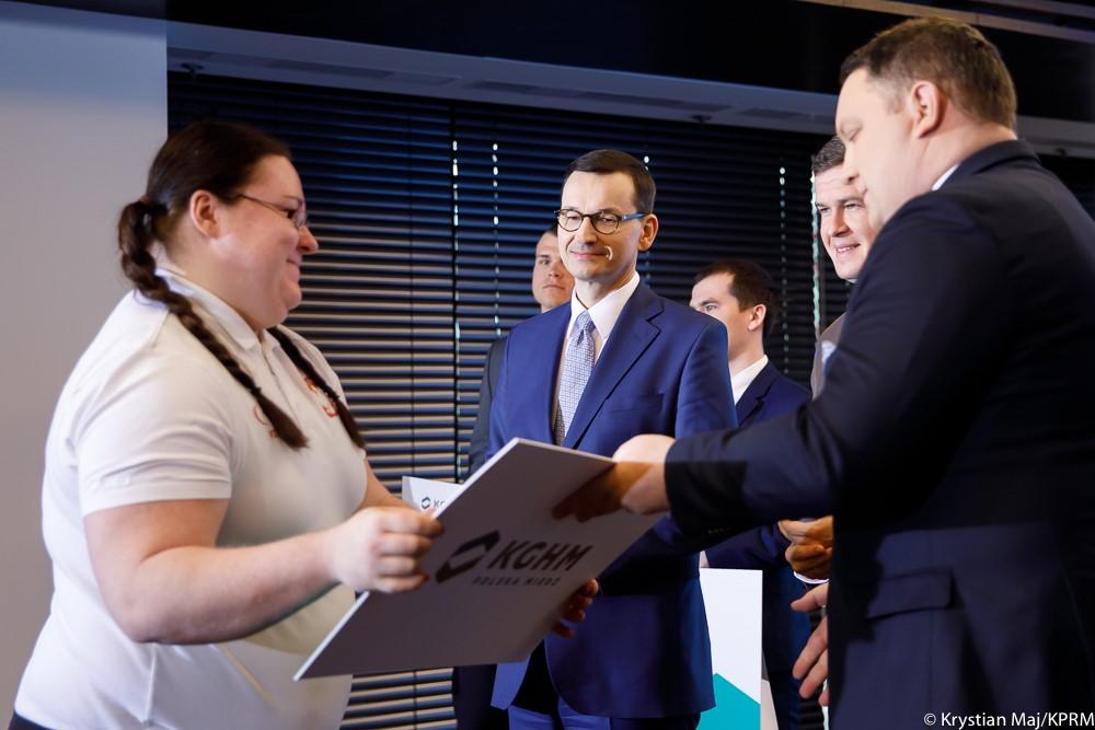 Prezes zarządu KGHM Marcin Chludziński wręcza czek stypendialny kobiecie, a z tyłu stoi premier Mateusz Morawiecki.
