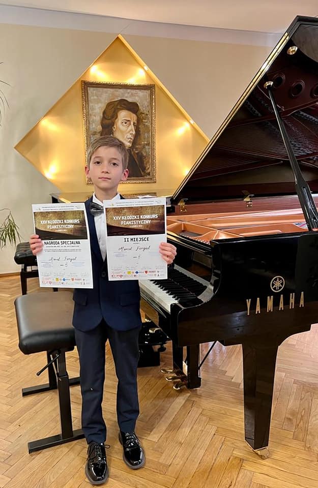 Na zdjęciu widoczny uśmiechnięty chłopiec, który w rękach trzyma dwa dyplomy. W tle stoi czarny fortepian firmy "Yamaha" 