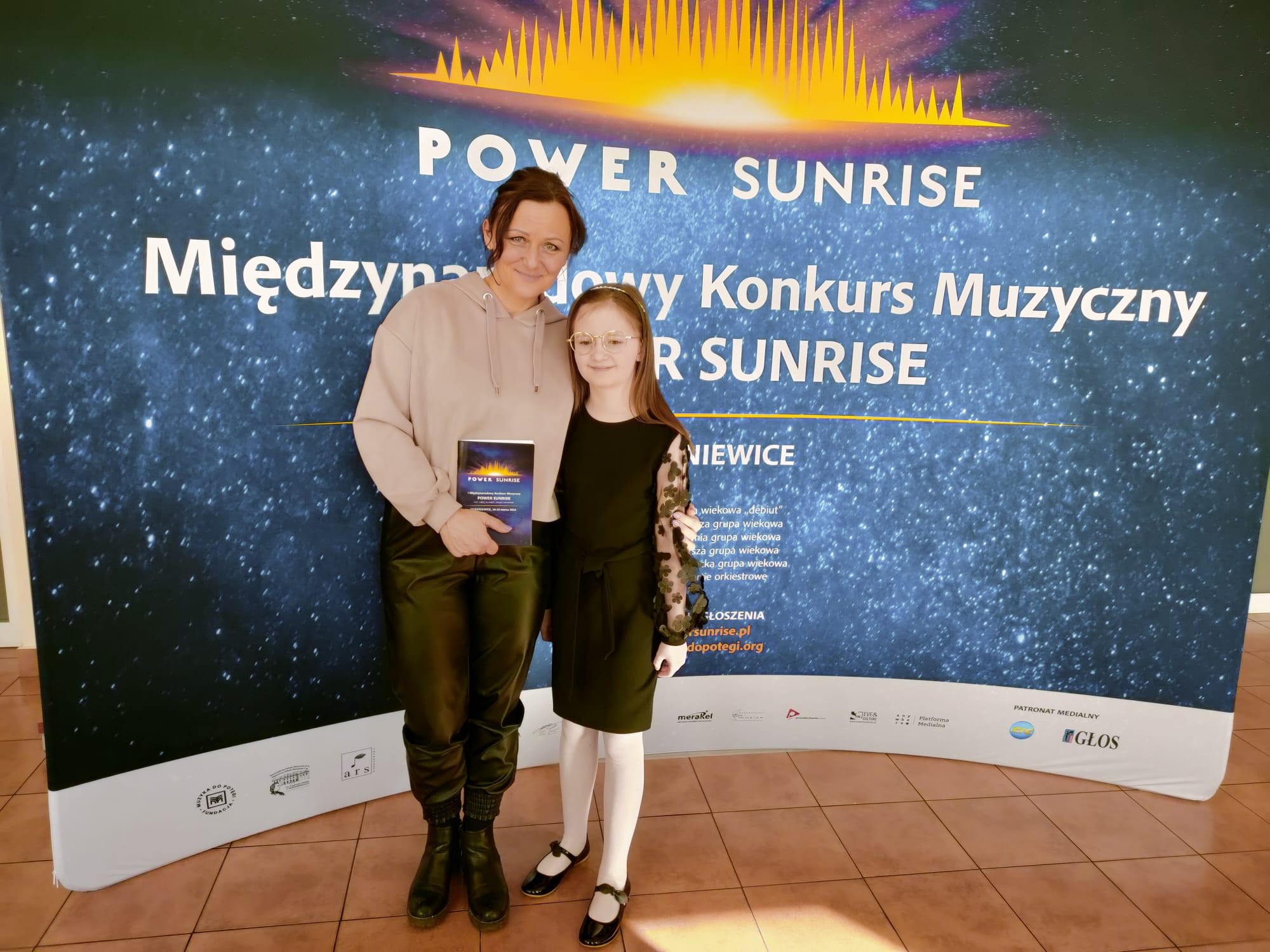 Uczennica i nauczycielka stoją uśmiechnięte na tle banera reklamowego konkursu