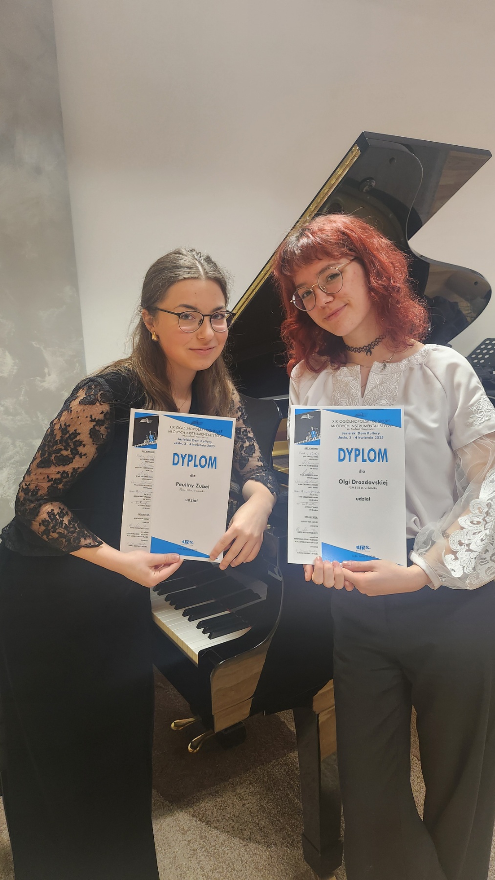 Paulina Zubel i Olga Drozdowska z dyplomami konkursu. W tle fortepian.