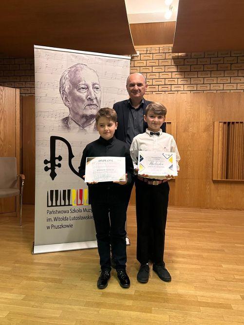 Dwóch uczniów trzymających dyplomy laureatów wraz z nauczycielem po środku.