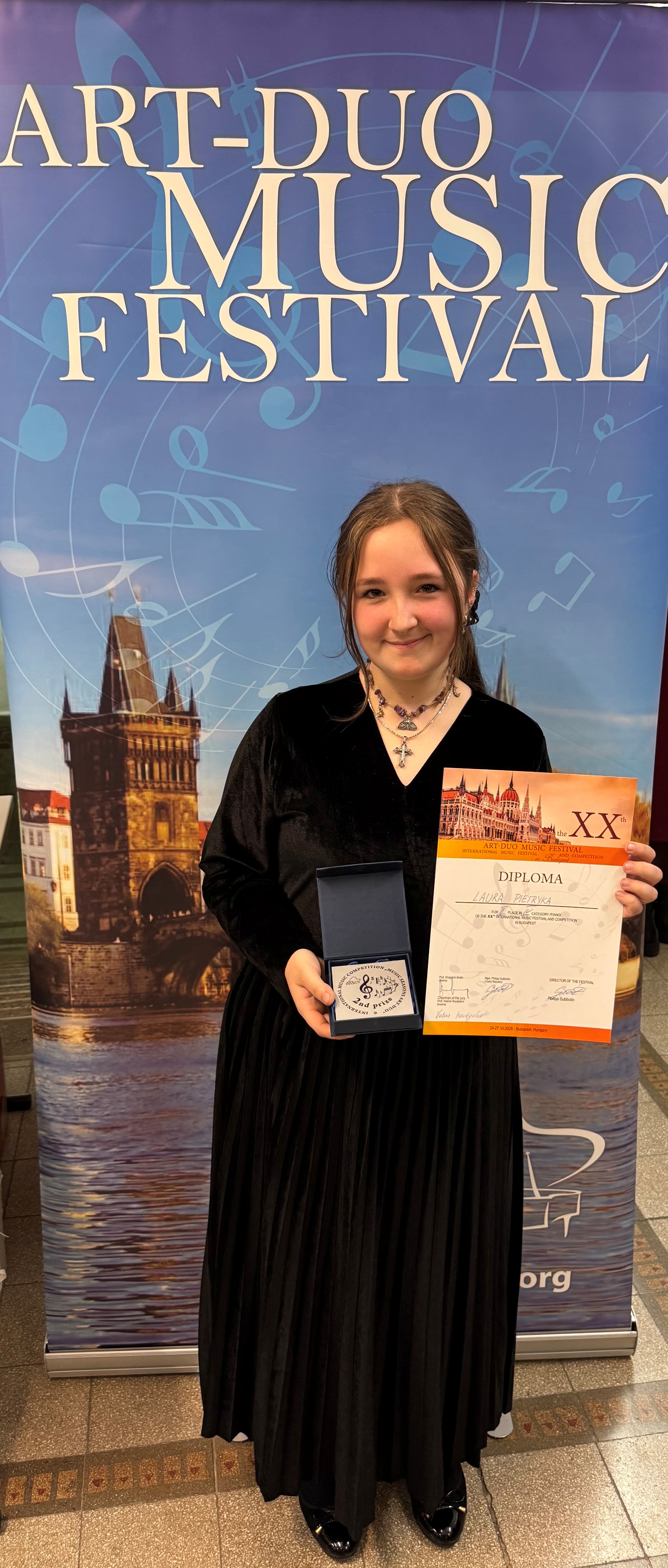 Laura Pietryka Art Duo 2025. Uczennice stojąca z dyplomem i medalem przed banerem reklamowym konkursu