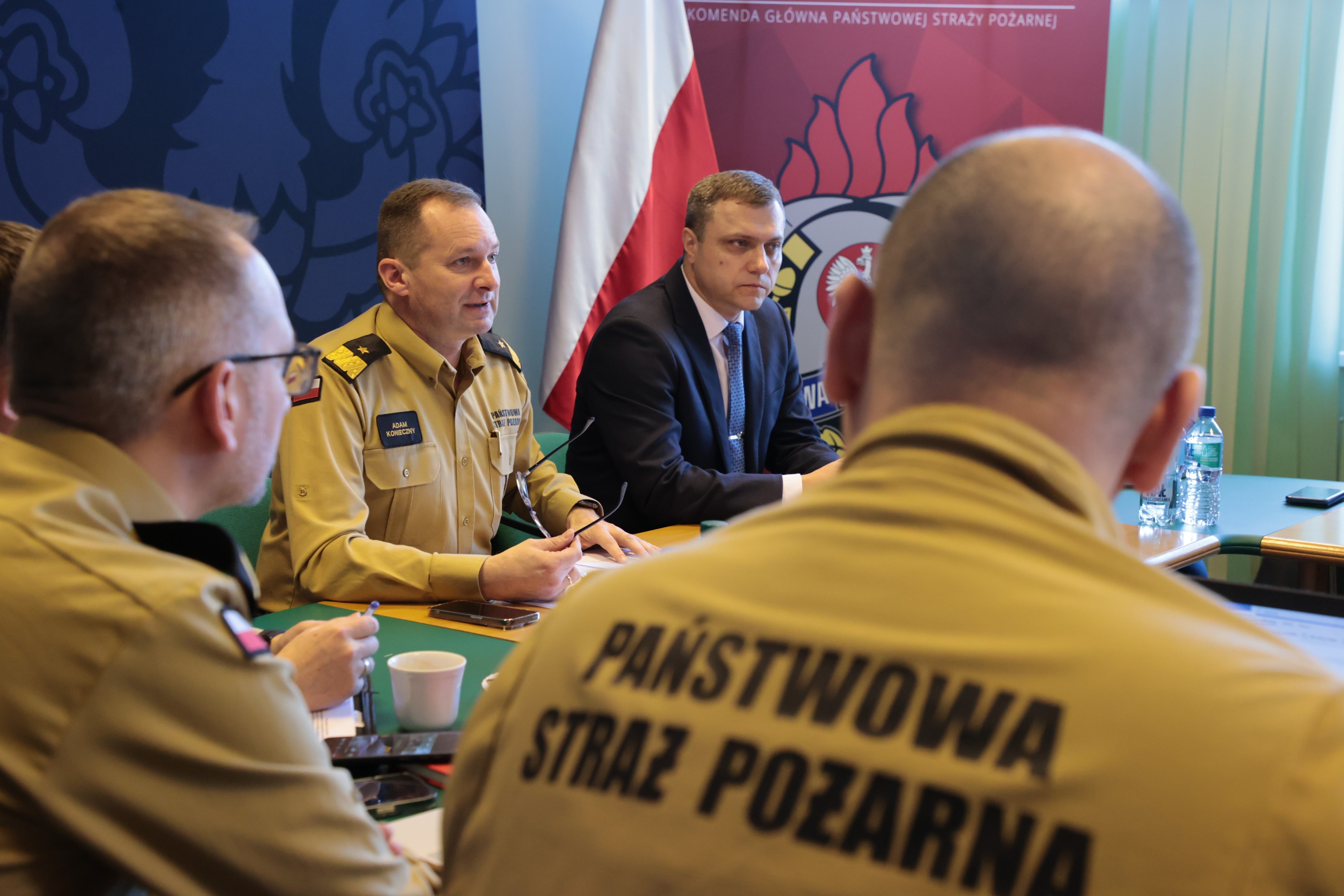 Dwóch strażaków siedzi podczas spotkania (widok z tyłu), między nimi widoczni zastępca komendanta głównego PSP wspólnie z przedstawicielem czeskiej delegacji siedzą przy stole prezydialnym.