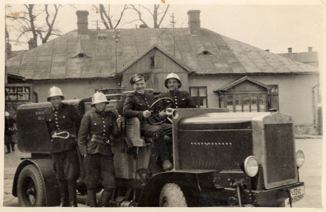 Samochód pożarniczy „beczkowóz” ZSP w Nowym Sączu z 1944 roku i czterech strażaków dwóch siedzi a dwóch stoi. W tle stary budynek straży pożarnej.