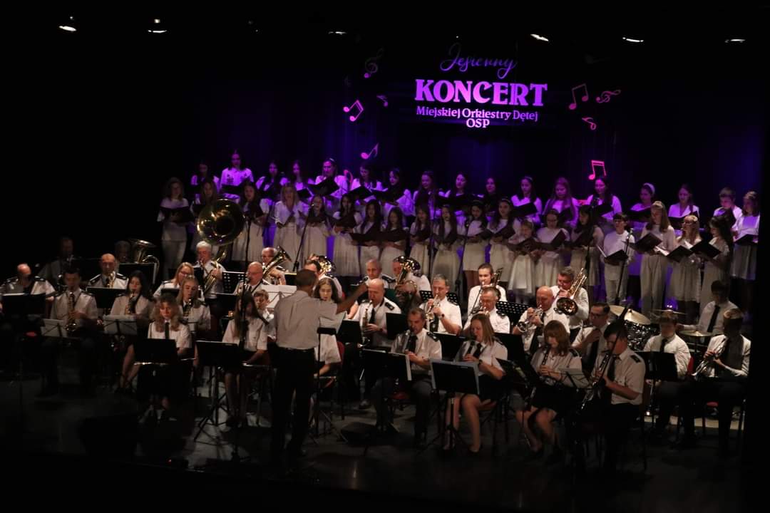 Na scenie znajduje się orkiestra dęta oraz chór dziecięcy ubrany na biało. Przed orkiestrą stoi dyrygent. 