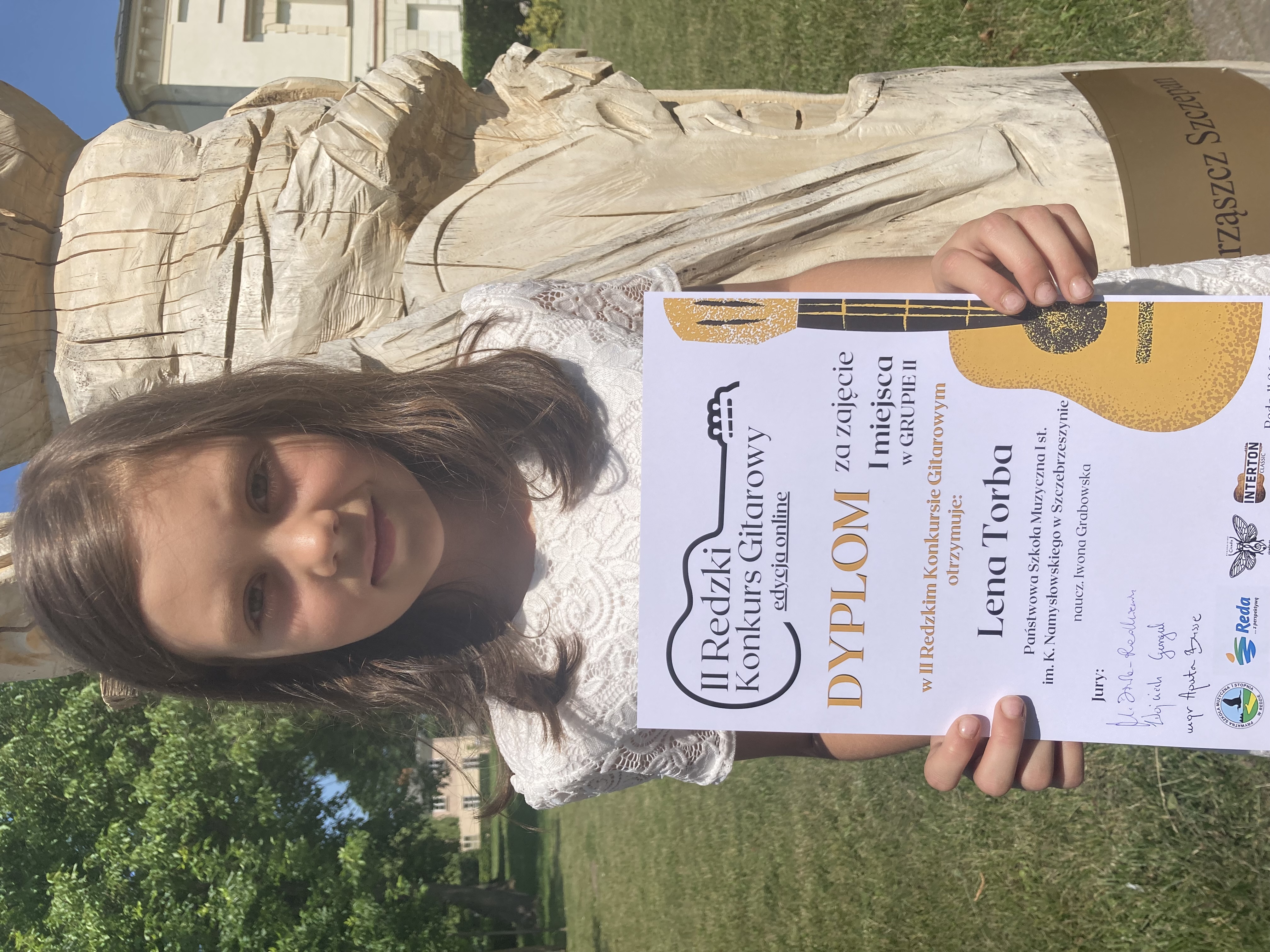 Laureatka II Redzkiego Konkursu Gitarowego (edycja online) Lena Torba. Trzyma dyplom z konkursu za zajęcie I miejsca. Stoi przed drewnianym chrząszczem Szczepanem przed nasza szkołą