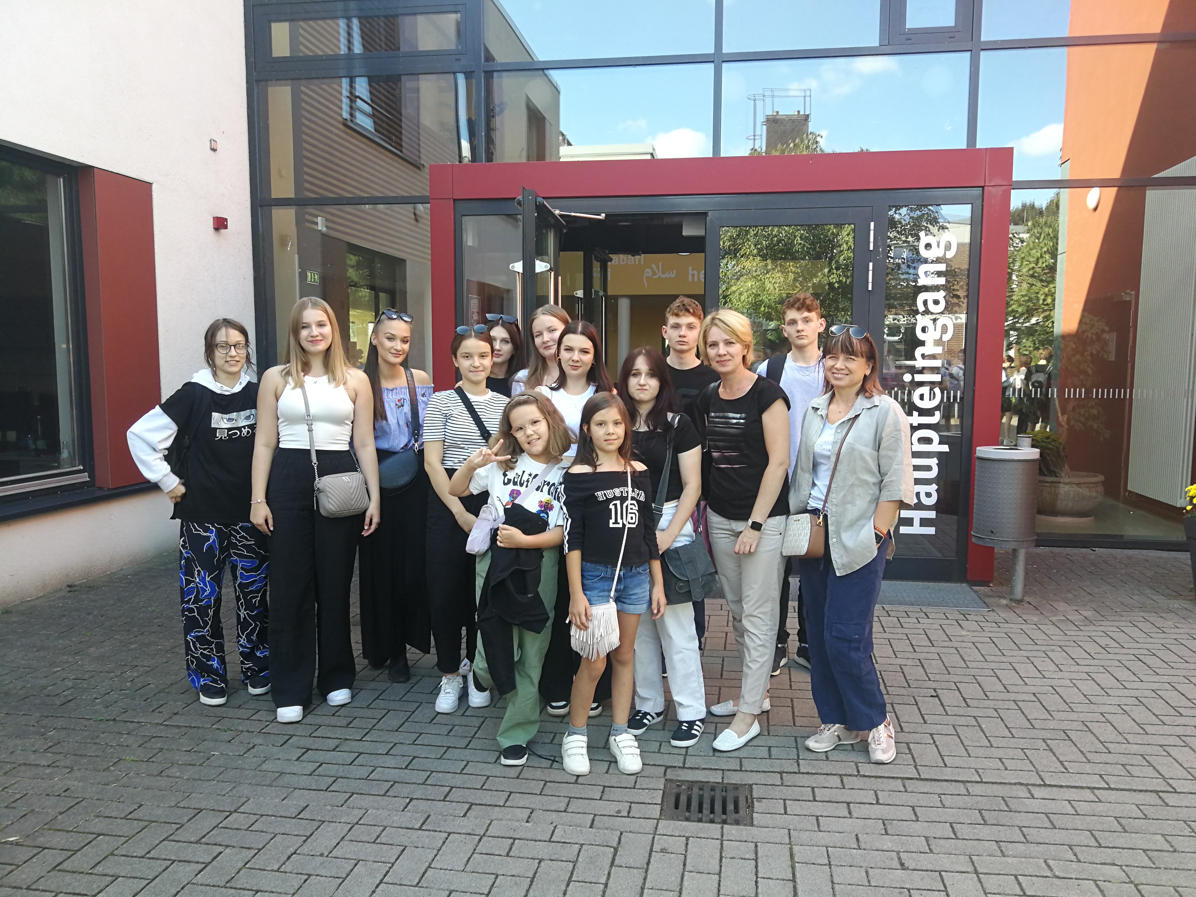 Zdjęcie przedstawia grupę uczniów i nauczycieli przed wejściem do budynku. Od lewej: Malwina Piwińska, Oliwia Bielecka, Amelia Pankiewicz, Gabriela Kościk, Kamila Wróblewska, Amelia Lewkowicz, Katarzyna Wiśniewska, Matylda Kędra, Lena Torba, Jagoda Piwińska, Mateusz Zieliński, Małgorzata Kawalec-Pyś, Radosław Zieliński, Iwona Grabowska