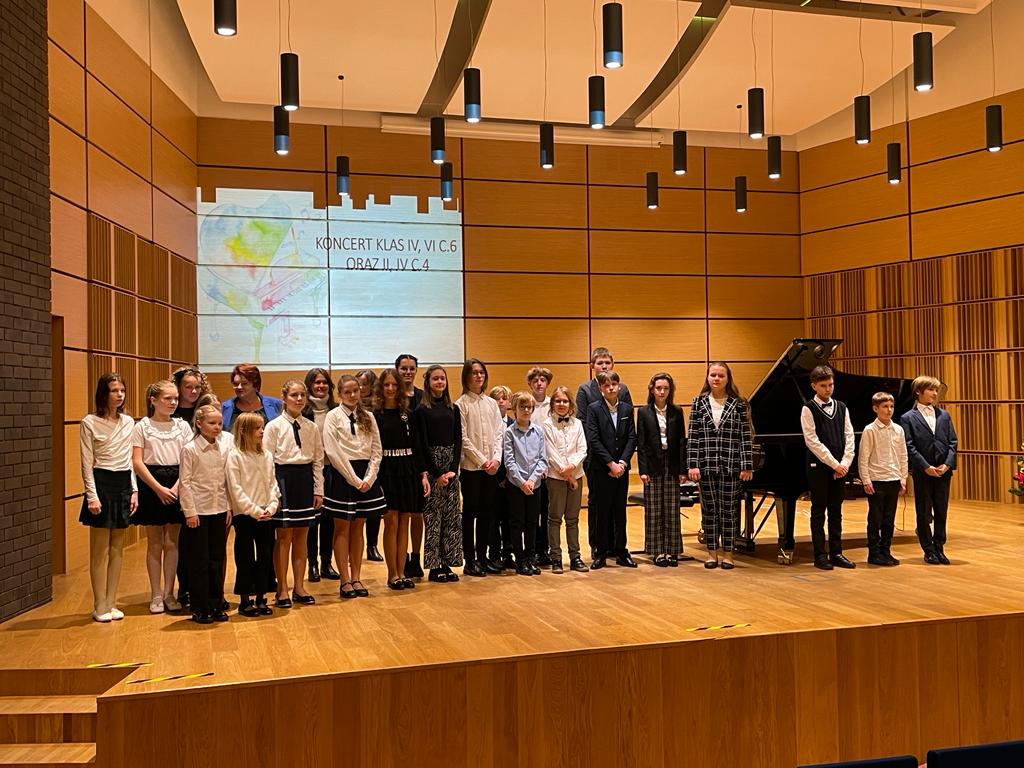 Uczniowie wraz z nauczycielami stojący w trzech rzędach na scenie sali koncertowej. Na górze widoczne wiszące lampy. Z tyłu wyświetlany na projektorze napis: "Koncert klas IV, VI c.6 oraz II i IV c.4".