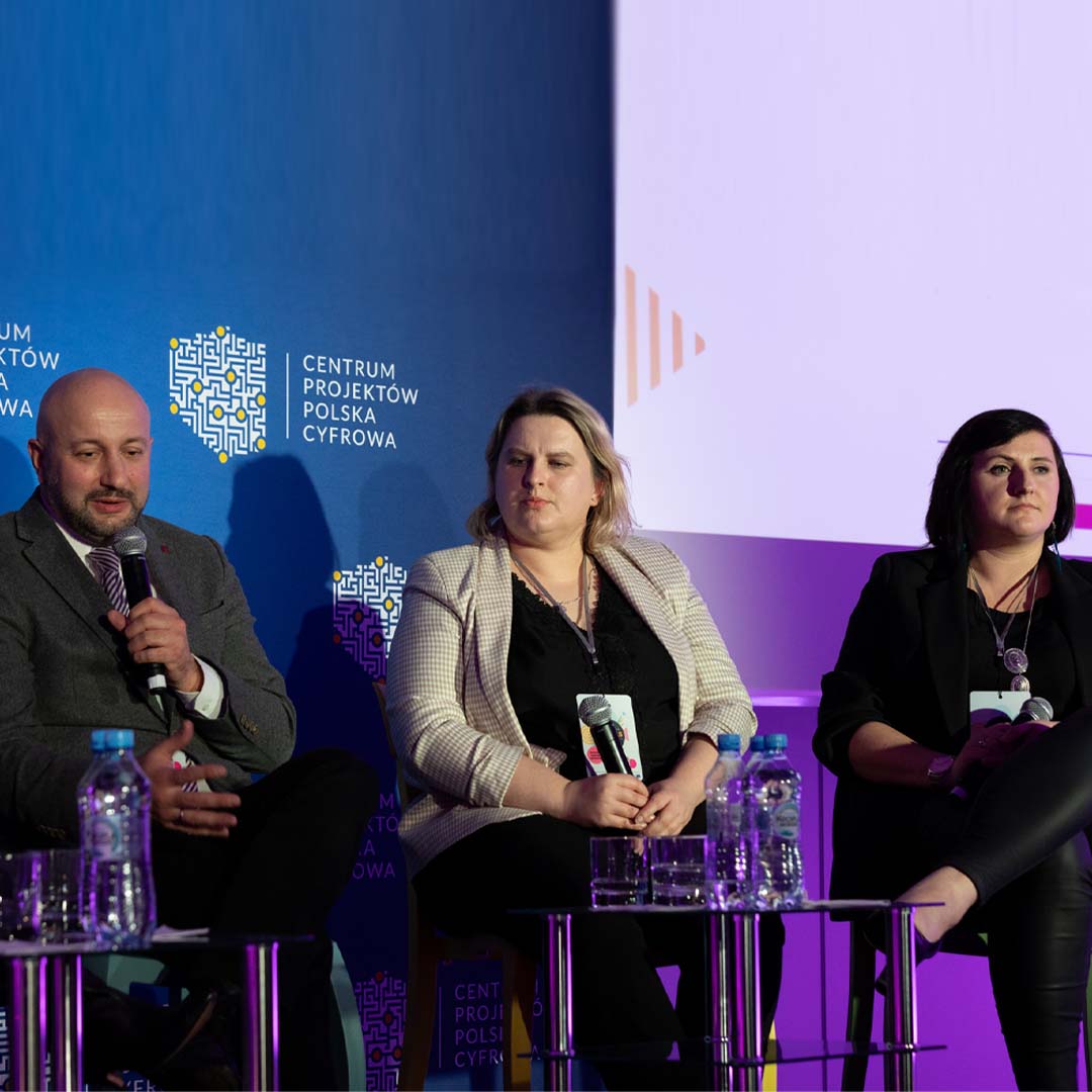 Pan Hubert Gęsiarz mówi do mikrofonu, Pani Maria Zdziech i Pani Katarzyna Jaszczak-Bukowska słuchają. Wszyscy siedzą na krzesłach na scenie.