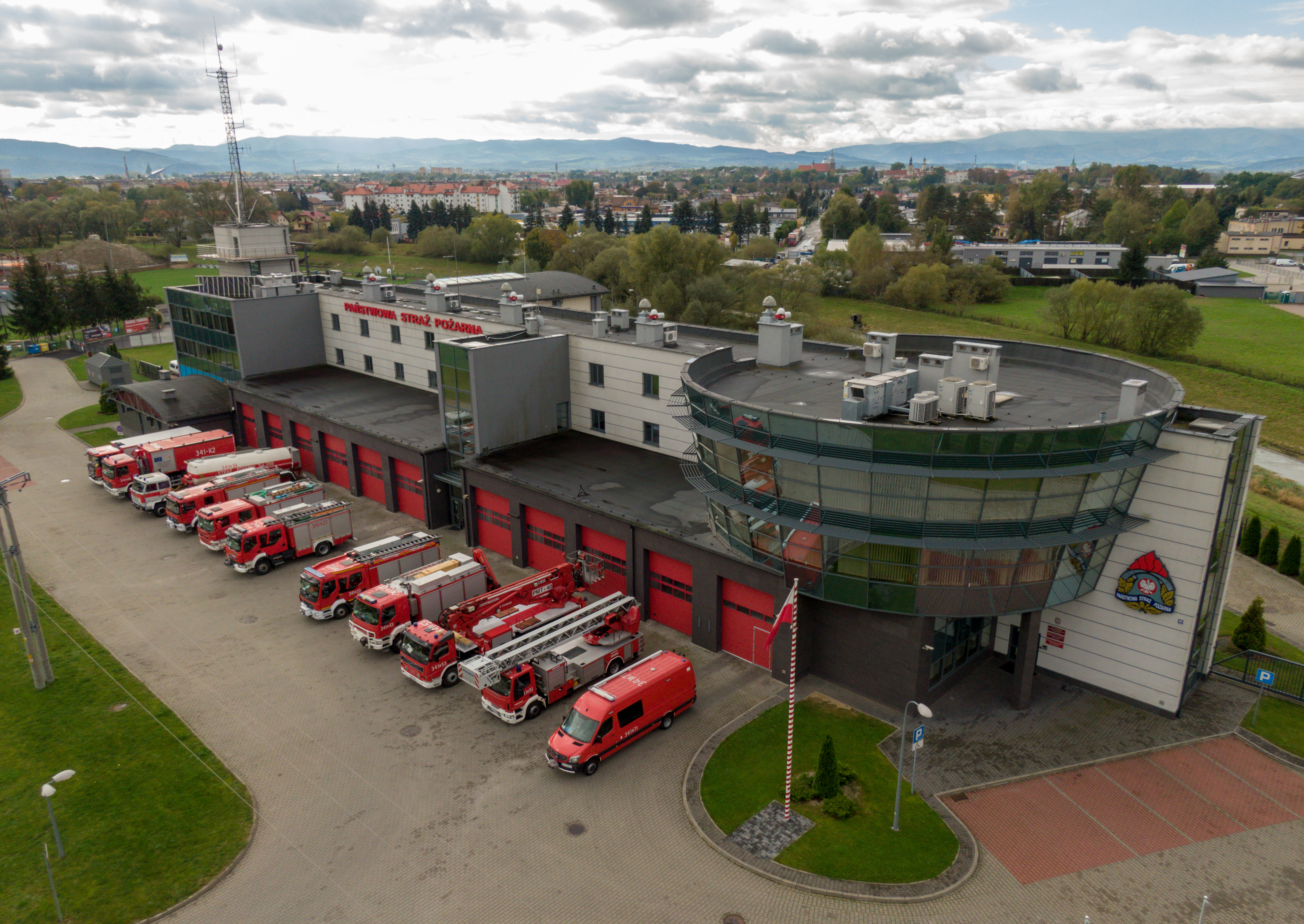 Widok nowej strażnica Komendy Miejskiej i JRG Nr 1 Nowy Sącz z października 2020. Przed budynkiem na placu zewnętrznym stoją samochody pożarnicze. W tle widać część Nowego Sącza oraz otaczające pasma górskie.