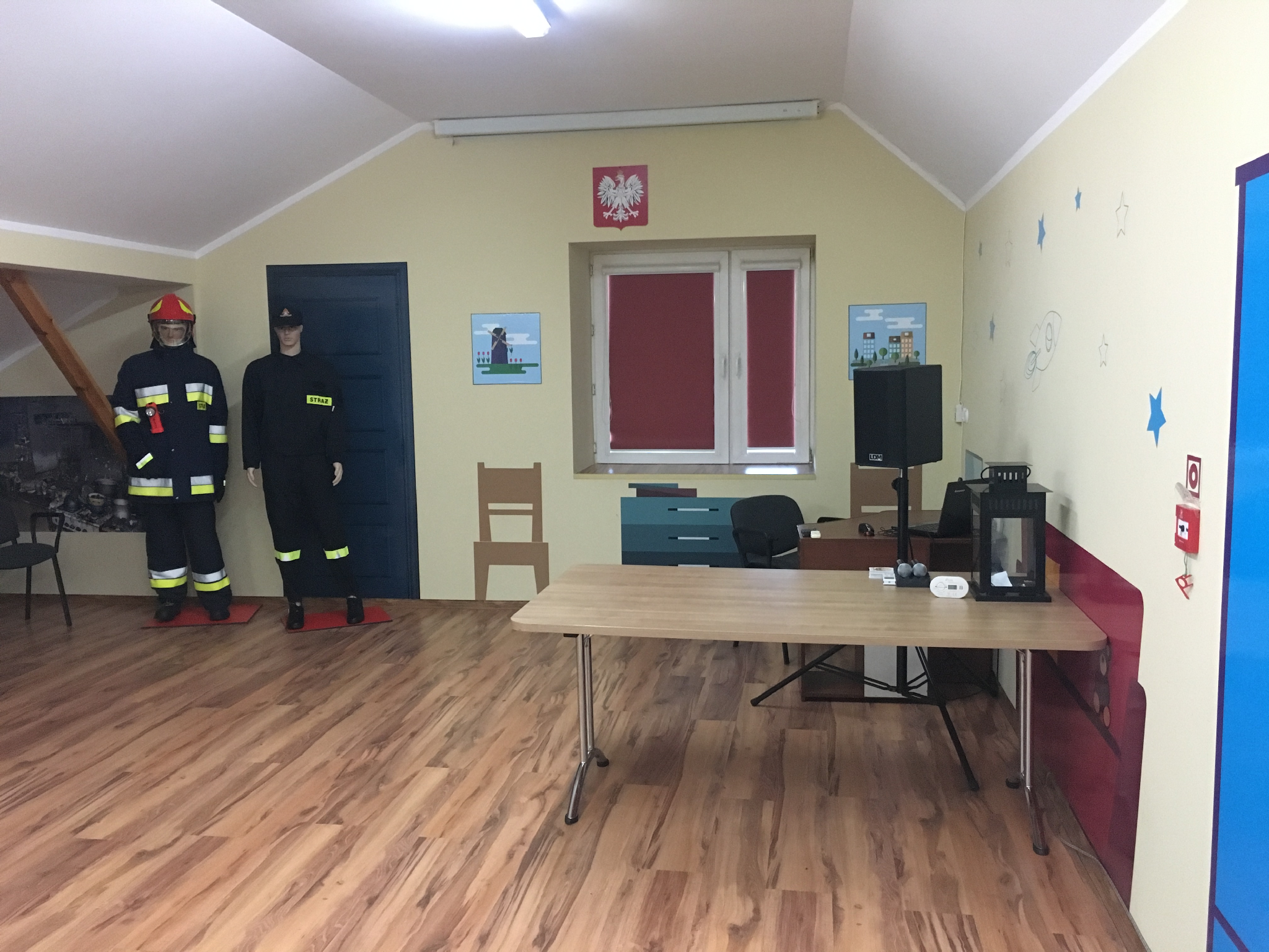 Zdjęcie przedstawia salę edukacyjną, po prawej stronie stanowisko do prowadzenia zajęć, po lewej 2 manekiny strażaka ubrane w mundur.