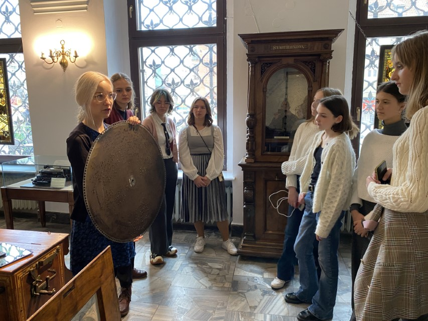 Zdjęcie kolorowe. Grupa uczniów w muzeum. Przewodniczka pokazuje miedzianą płytę.