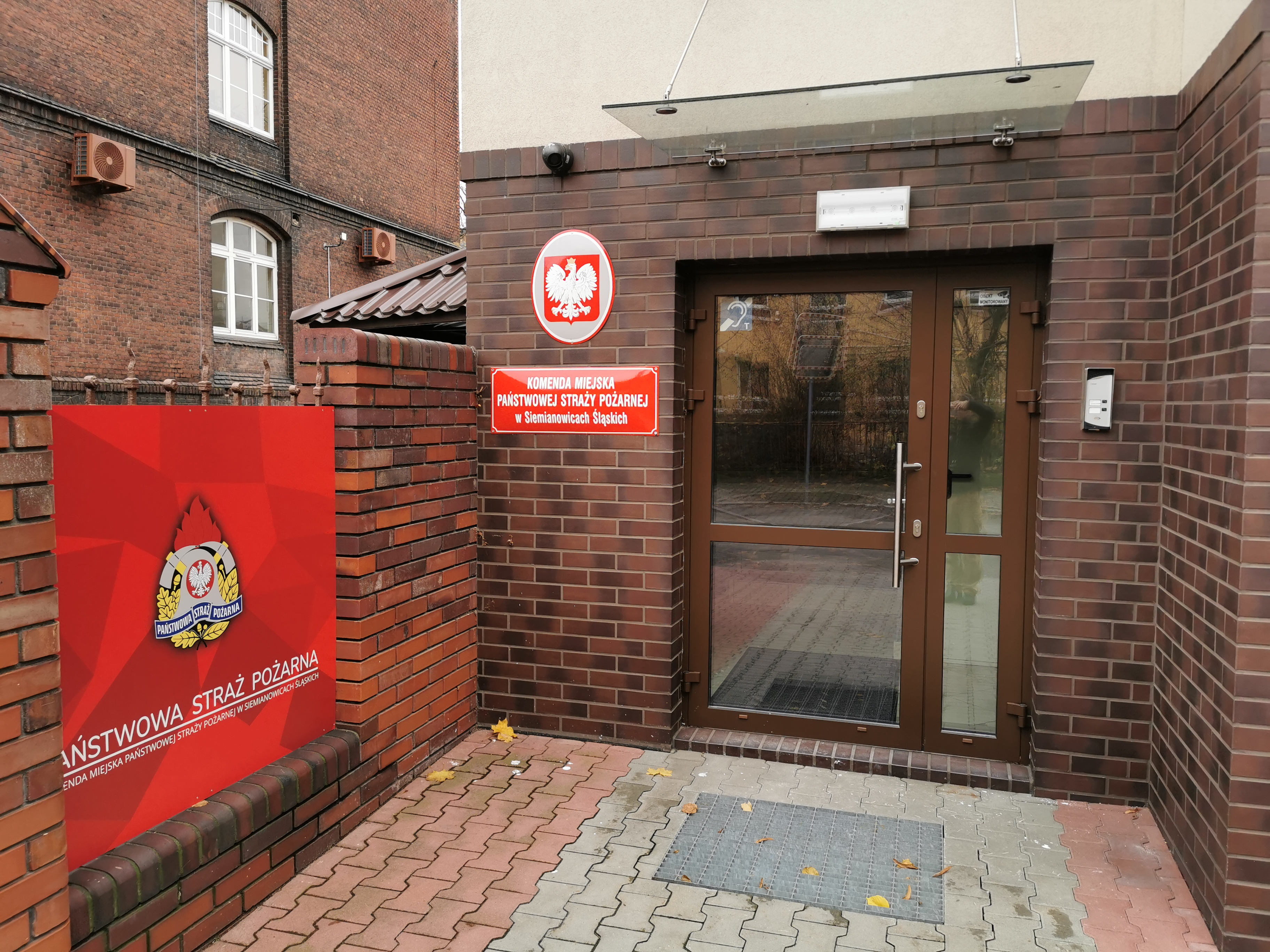 Na zdjęciu widoczne jest główne wejście do budynku Komendy Miejskiej Państwowej Straży Pożarnej w Siemianowicach Śląskich. Wejście wykonane jest z czerwonej cegły, drzwi są przeszklone z ramą w kolorze brązowym. Obok drzwi znajdują się tablice informacyjne: • Czerwona tablica z białym godłem Polski, poniżej napis: „KOMENDA MIEJSKA PAŃSTWOWEJ STRAŻY POŻARNEJ w Siemianowicach Śląskich” • Po lewej stronie znajduje się większy, czerwony szyld z logo Państwowej Straży Pożarnej i napisem „PAŃSTWOWA STRAŻ POŻARNA KOMENDA MIEJSKA PSP W Siemianowicach Śląskich”.