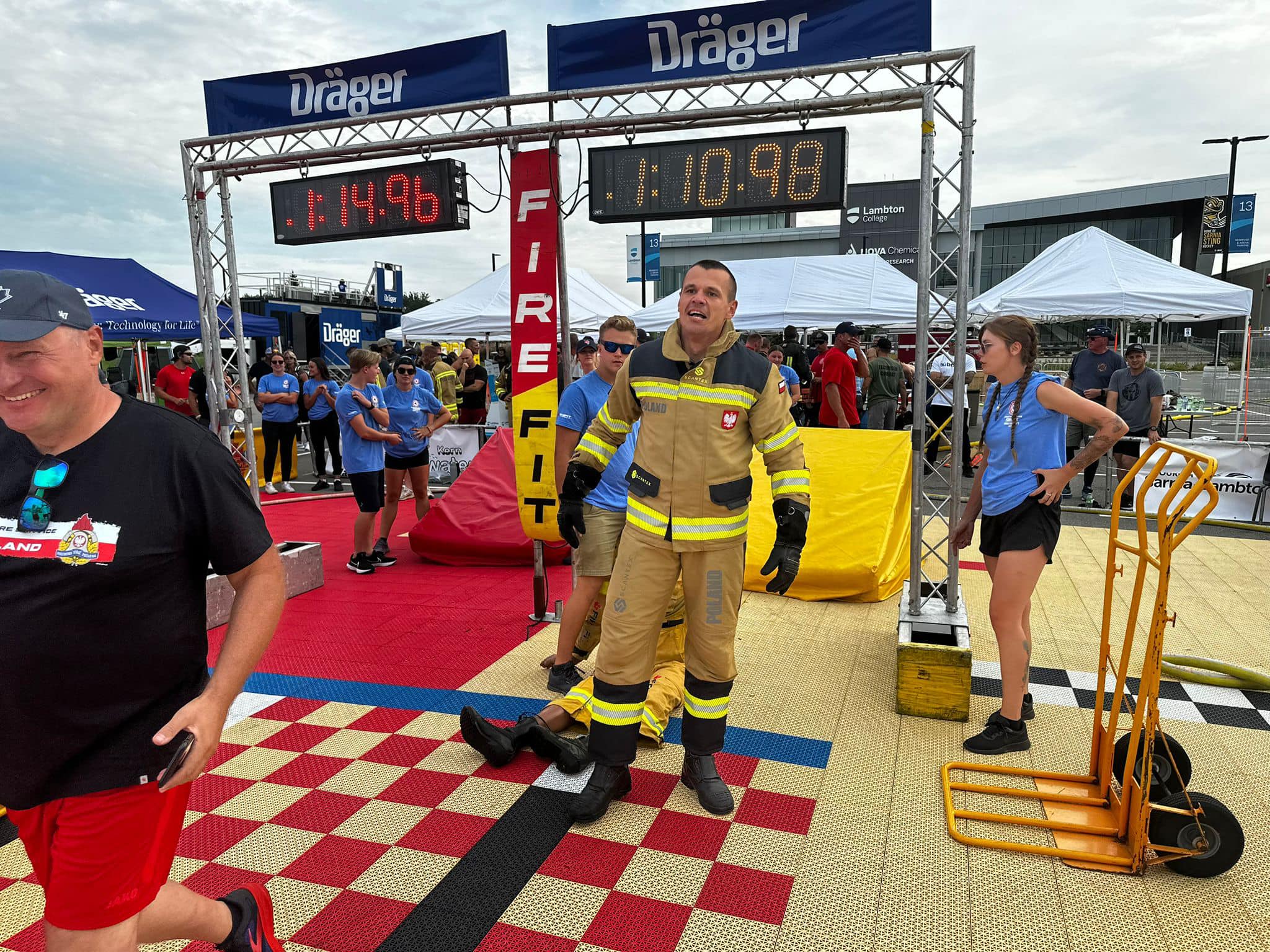 Zawodnik w ubraniu strażackim stoi na macie sportowej za nim tablica sportowa z wyświetlonym wynikiem 1;14.96