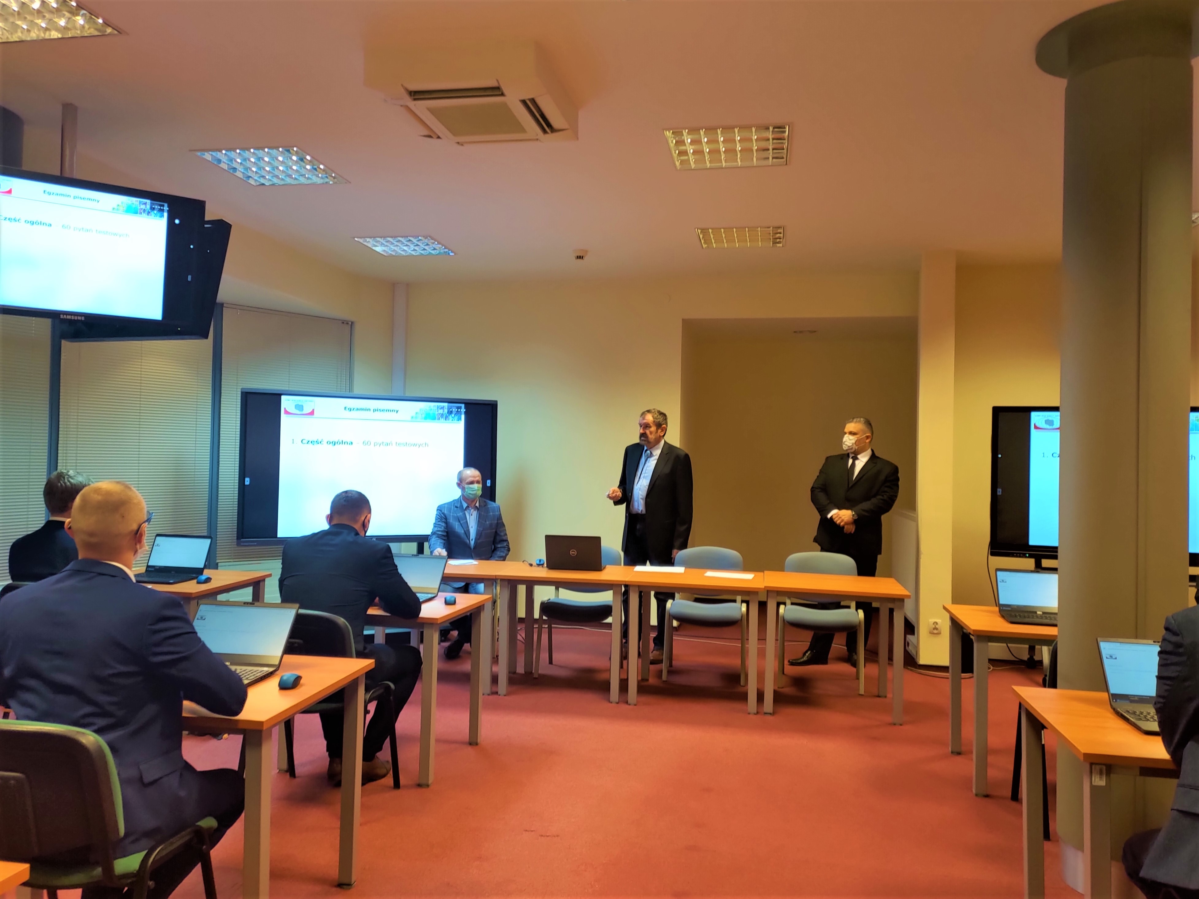 Zdjęcie przedstawia salę egzaminacyjną z członkami Komisji egzaminacyjnej i osobami przystępującymi do egzaminu siedzącymi pojedynczo przed laptopami