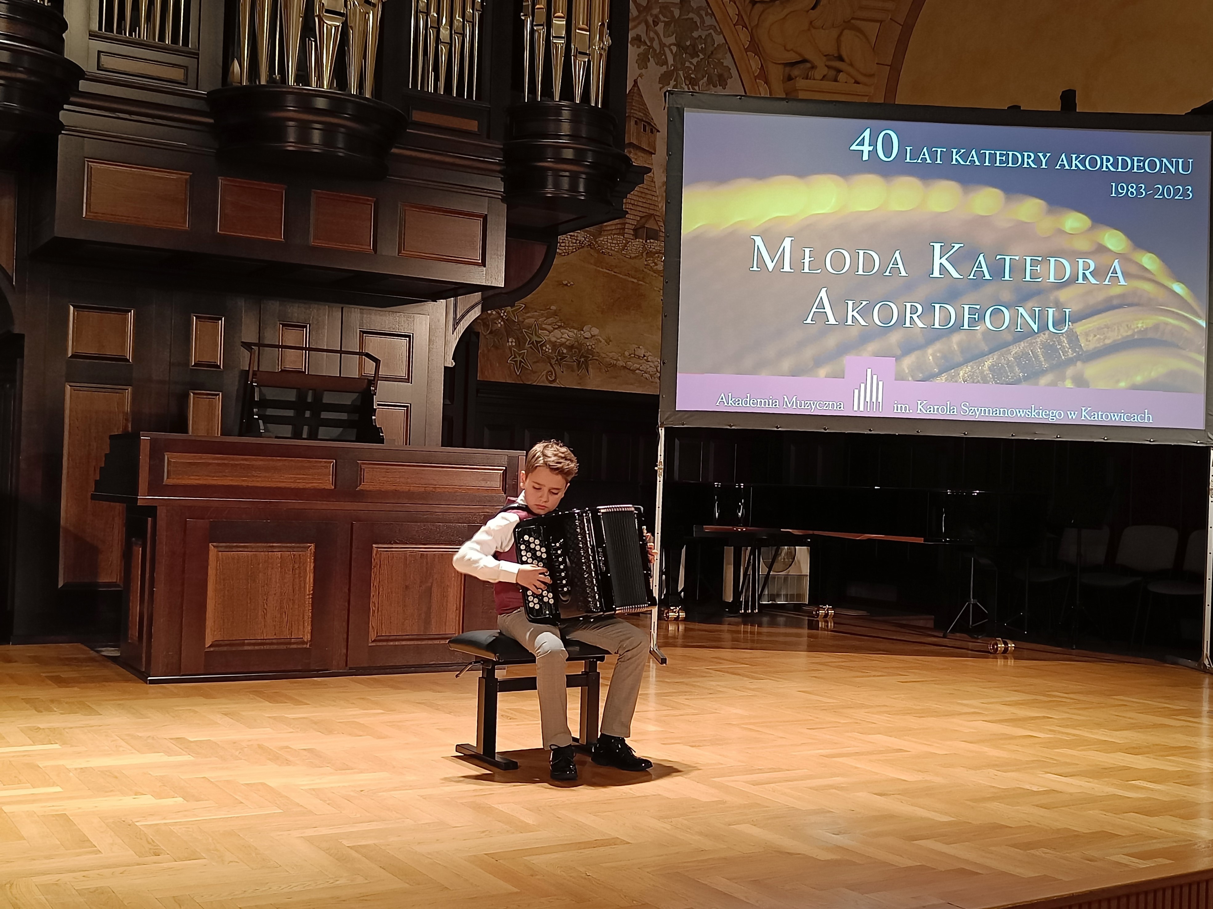 Uczeń siedzący na czarnym krześle, grający na akordeonie. Z tyłu widoczne organy w kolorze brązowym. Po prawej stronie rzutnik z wyświetlonym slajdem: 40 lat katedry akordeonu 1983-2023, Młoda Katedra Akordeonu, Akademia Muzyczna im. Karola Szymanowskiego w Katowicach.