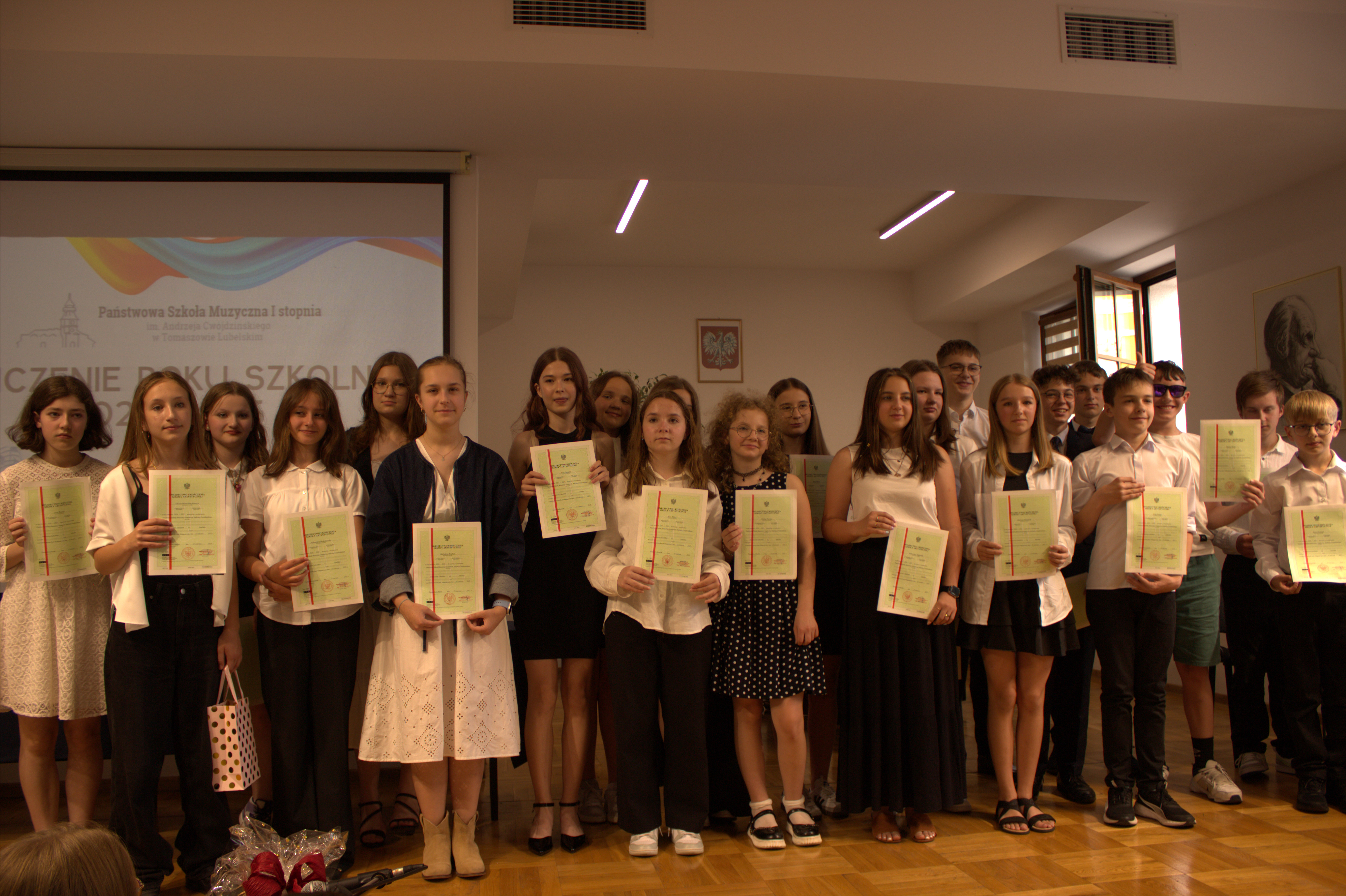 absolwenci trzymają w dłoniach świadectwa i pozują do zdjęcia grupowego na estradzie sali koncertowej