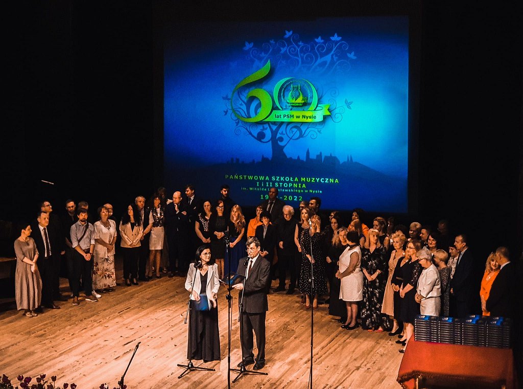 Zdjęcie. Jubileusz 60-lecia PSM w Nysie część oficjalna - 13 maja 2022 - Nyski Dom Kultury. na scenie z przodu stoją wicedyrektor Beata Toczek i dyrektor Janusz Dąbrowski. W głębi nauczyciele i pracownicy szkoły. W tle logo jubileuszu wyświetlone od tyłu na wiszącym ekranie. Kliknięcie na zdjęcie przeniesie do prezentacji cyklu zdjęć z koncertu.
