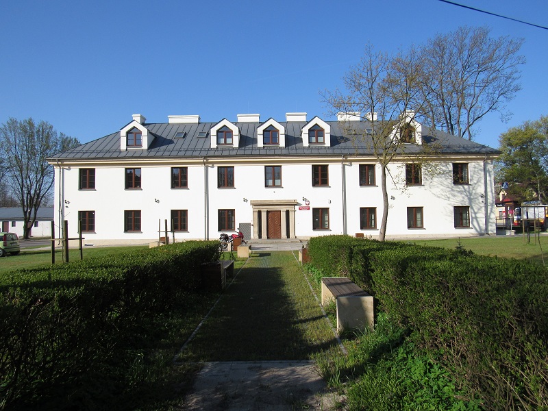 Zdjęcie przedstawia biały dwupiętrowy budynek. Drugie piętro znajduje się w ciemno grafitowym dachu. W tle niebieskie niebo. Do szkoły prowadzi ozdobne drewniane drzwi otoczone kamienną ornamentyką z filarami. Przed szkołą zielony trawnik i żywopłot. 