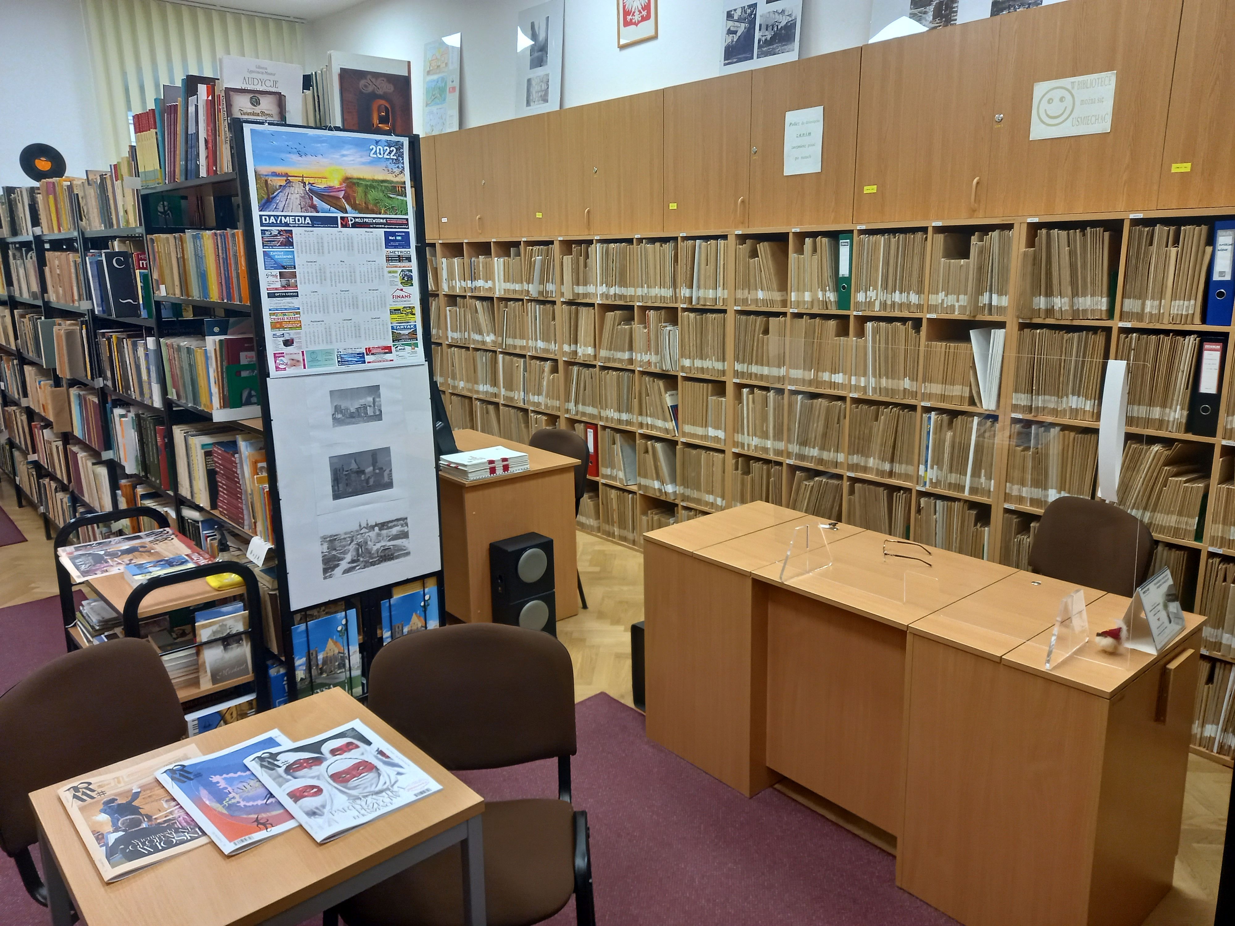 Zdjęcie. Wnętrze biblioteki. Na pierwszym planie stolik z czterema krzesełkami i biurko. Na stoliku leżą czasopisma Ruch Muzyczny. W środku regały z książkami i nutami oraz szafki z płytami CD.
