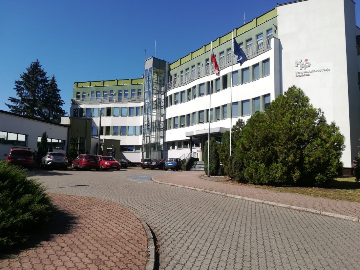 trzypiętrowy budynek z napisem po jednej stronie: KAS Krajowa Administracja Skarbowa, przed budynkiem miejsca parkingowe i droga dojazdowa
