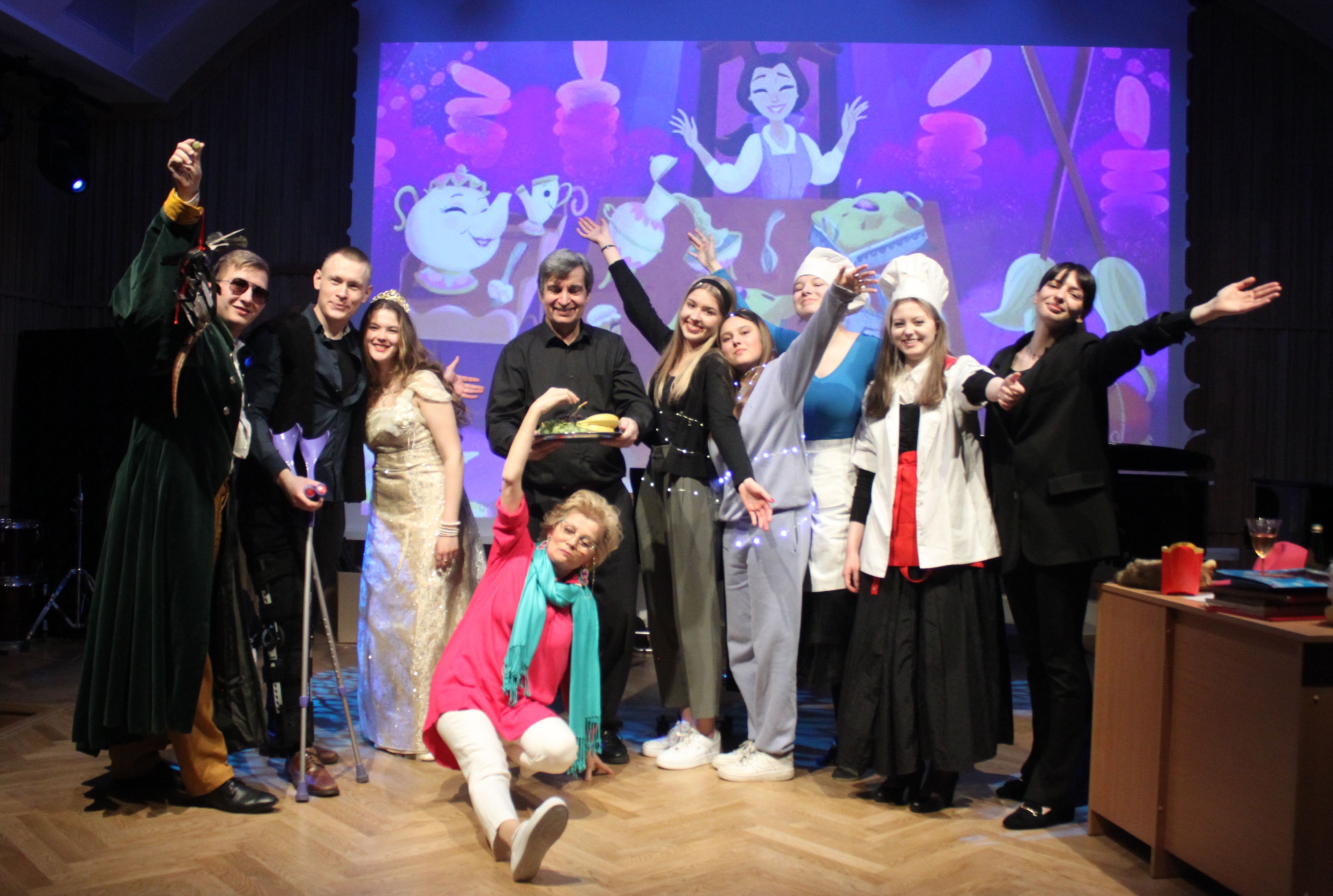 Grafika - zdjęcie. Grupa aktorów po spektaklu na estradzie pozuje do wspólnego zdjęcia. W tle fragment scenografii wyświetlony na ekranie.