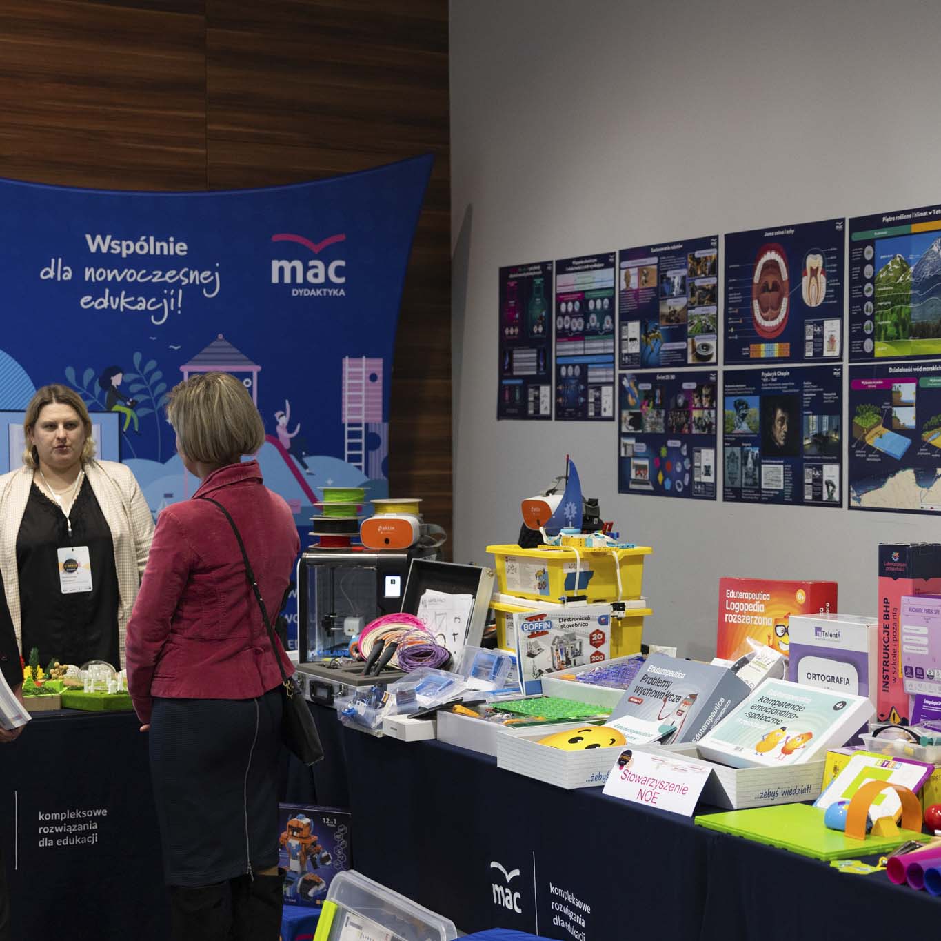 Przy stoisku MAC stoją 3 kobiety. Na stoisku leżą klocki, materiały dydaktyczne, drukarka 3d.