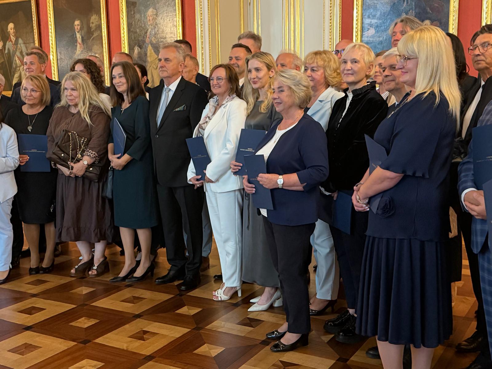 Grupowe zdjęcie przedstawiające grupę osób w eleganckich strojach, stojących w dwóch rzędach w wytwornym, zabytkowym wnętrzu. Kobiety mają na sobie sukienki lub żakiety, a mężczyźni garnitury, najczęściej w kolorach granatowym, czarnym lub szarym. Większość z nich trzyma w rękach granatowe teczki. Uczestniczą w uroczystości wręczenia. Niektórzy patrzą prosto w obiektyw i się uśmiechają, inni rozmawiają między sobą. Tło zdjęcia stanowi ściana obwieszona licznymi portretami w bogato zdobionych, złoconych ramach. Drewniana podłoga jest ułożona w geometryczny wzór. Światło jest naturalne i rozproszone, oświetlając grupę równomiernie.