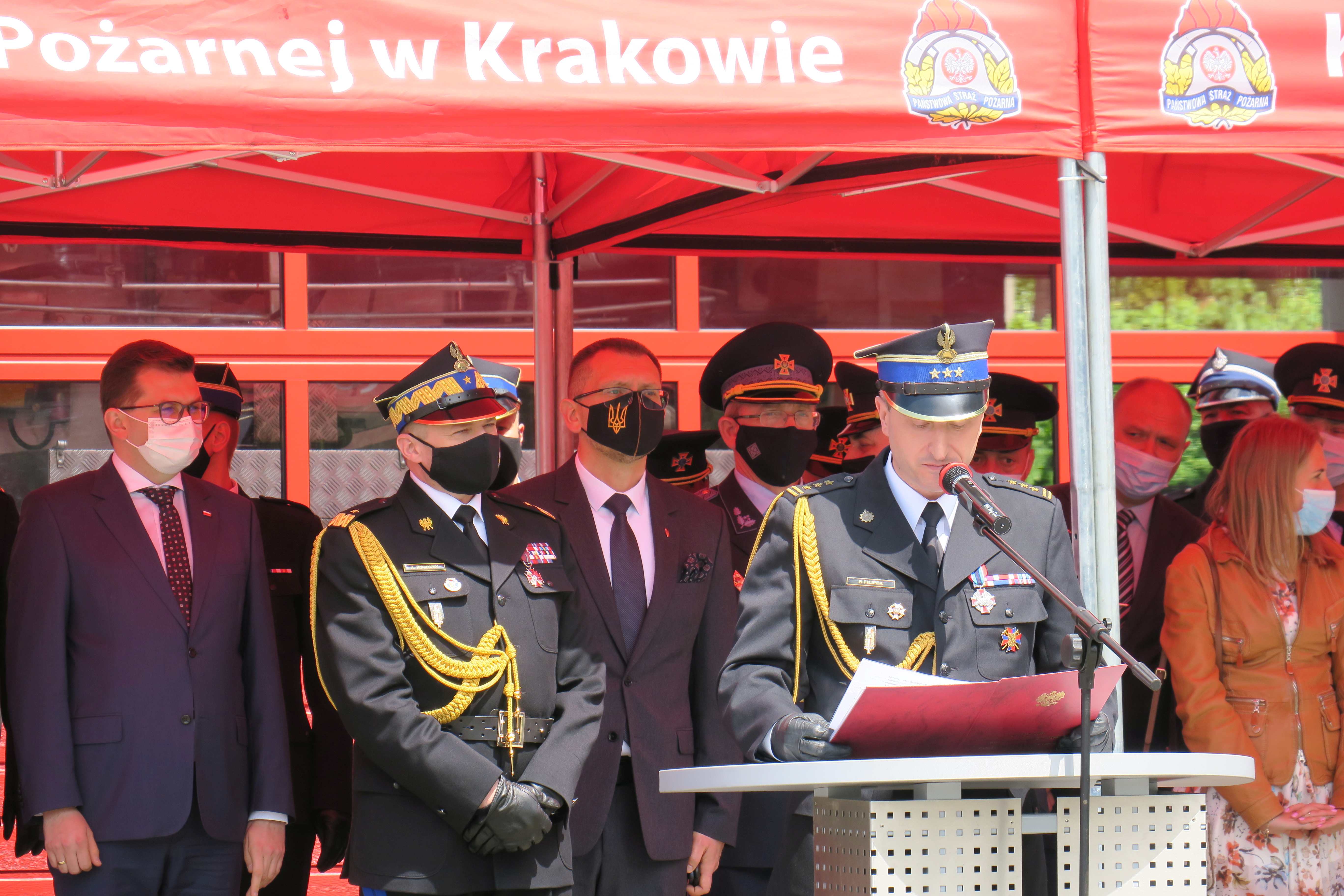 Trybuna honorowa na której pod czerwonym baldachimem stoją strażacy w mundurach galowych. Na pierwszym planie przemiawia małopolski komendant wojewódzki PSP.