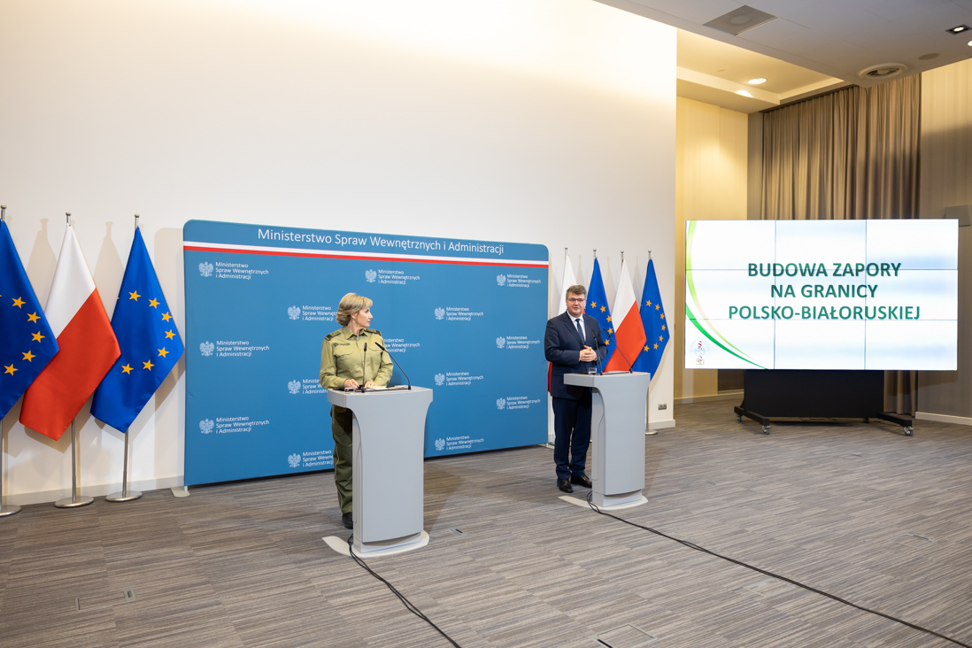 Na zdjęciu widać wiceministra Macieja Wąsika i gen. bryg. SG Wioletę Gorzkowską stojących za mównicami w trakcie konferencji prasowej. W tle widać ściankę MSWiA, flagi Polski i UE i ekran z wyświetloną prezentacją.